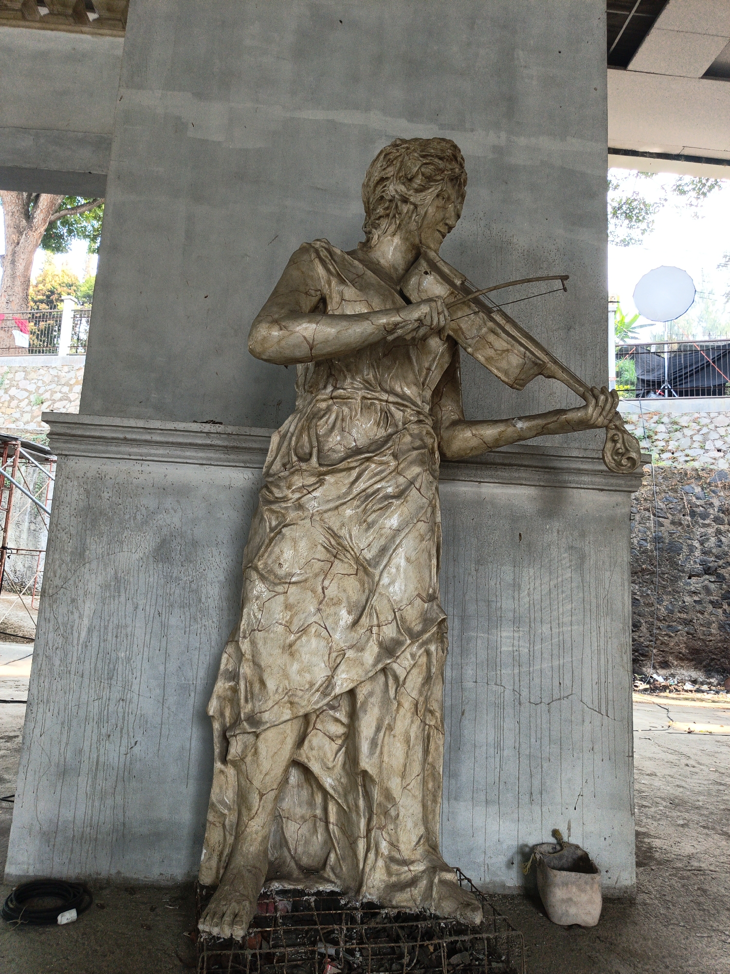 A full-body, weathered statue of a woman playing a violin stands against a concrete pillar. The statue is light brown with dark cracks running through its surface, suggesting age or a deliberate artistic effect. She is dressed in a flowing, draped gown. Her hair is styled in curls, and her face is turned to her right as she looks down at the violin. Her left hand holds the neck of the violin, and her right hand draws the bow across the strings. The statue rests on a metal grate at its base. To the left of the statue, a coiled black cable lies on the ground. To the right, a weathered, open container sits on the ground. Behind the statue, a concrete wall with streaks of dark discoloration is visible. In the background, there is a glimpse of trees, a fence, and a rocky slope. Further in the background, some scaffolding, a white disc, and a dark structure are visible. The scene appears to be outdoors in an unfinished or construction-like environment.