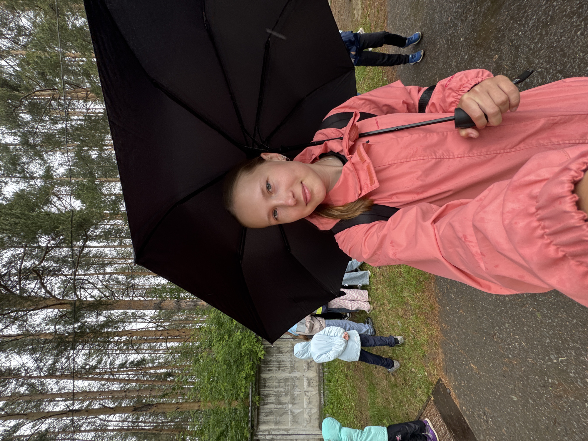 A young girl is depicted in a portrait-style shot, holding a large, open black umbrella outdoors, likely in Chaikovsky, Russia. She is positioned towards the left of the frame, looking directly at the viewer with a calm expression and a hint of a smile. She wears a coral-pink jacket, which appears to be waterproof, with "Outventure" faintly visible on the shoulder area. Her hair is light in color, possibly blonde or light brown.
The setting is an outdoor environment, characterized by a paved, wet path or road in the foreground and midground, indicating recent rainfall. To the left of the path, there is a stretch of green grass, behind which stands a low, weathered concrete wall. Beyond the wall, a dense line of tall pine trees with distinctive light-colored trunks and green needles forms the backdrop, suggesting a forested or park-like area.
Several other individuals are partially visible in the background, standing on the grass or path, some wearing jackets, further suggesting an outdoor activity or gathering. One person in the midground wears a light blue hooded jacket. The presence of the umbrella and the wet surfaces strongly imply that it is either raining or has recently rained. The sky, partially visible through the pine branches, appears overcast, consistent with diffused daylight. The overall atmosphere suggests a casual outdoor outing despite the damp weather.