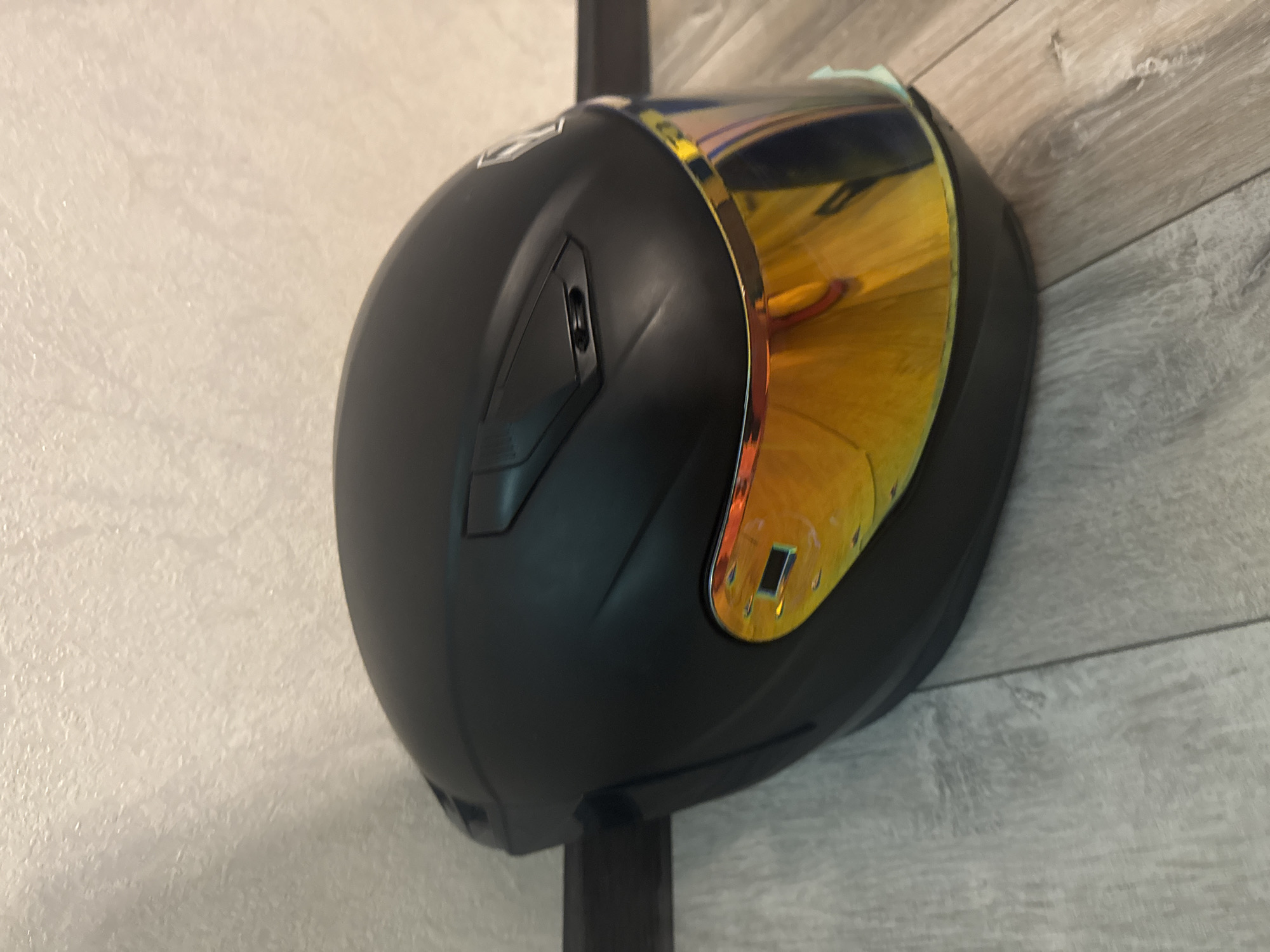 A black motorcycle helmet with a reflective gold visor is placed on a light-colored wooden floor against a textured off-white wall. The helmet is angled slightly, showing the top and right side. The visor is the most prominent feature, reflecting light and showing a distorted image of the surroundings. There are subtle vents and a brand logo on the top of the helmet. The background wall has a rough, textured finish. The floorboards are laid horizontally and have a wood grain pattern. The lighting appears to be artificial and casts soft shadows. There is a dark horizontal line along the base of the wall, possibly a baseboard or a shadow. There are no people visible in the image. The scene appears to be indoors, in a domestic setting.