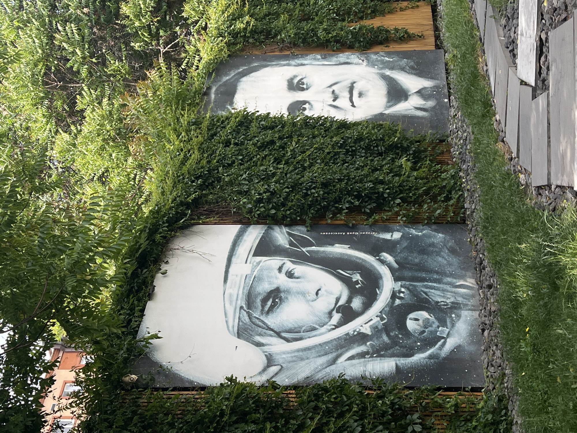 The image shows two portraits displayed outdoors in Unknown, Unknown. The lower portrait depicts a person wearing a space helmet, while the upper portrait shows a person in a suit. Both portraits are partially obscured by greenery and vines. A walkway of stone slabs and gravel runs alongside the portraits. The environment appears to be an urban garden or park.