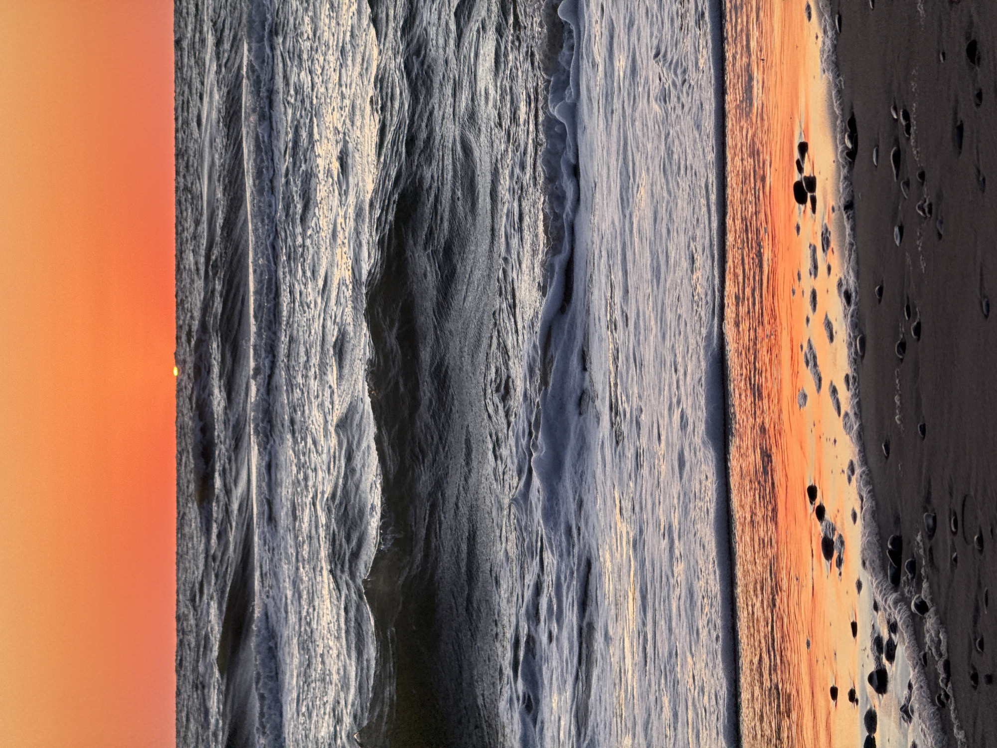 The image captures a serene sunset over the Baltic Sea at Ventspils, Latvia. The sky is a vibrant gradient of orange and red, with the sun, a small, glowing orb, just touching the horizon. The sea is a deep, dark blue, with gentle waves rolling towards the shore, creating white foam as they break. The wet sand on the beach reflects the warm colors of the sunset, and scattered pebbles dot the shoreline. The scene is tranquil and natural, with no people or man-made structures visible. The photograph is taken at eye-level, looking out towards the sea.