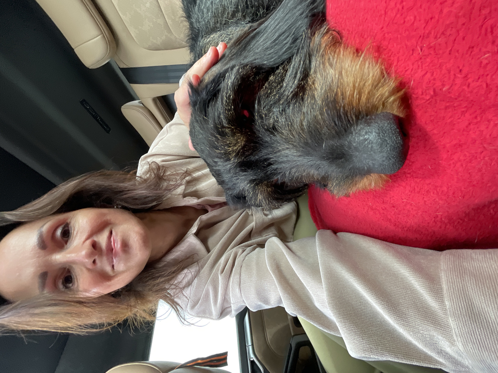 A woman and a dog are in the back seat of a car, possibly in Solov'evka, Russia. The woman is in the foreground, facing the camera and smiling. She has long, dark brown hair and is wearing a light-colored shirt. Her left hand is resting on the dog's head. The dog is a wire-haired dachshund with black and brown fur. It is lying down on a red blanket in the woman's lap. The car's interior is visible in the background, with beige leather seats. The lighting is natural, suggesting it is daytime.
