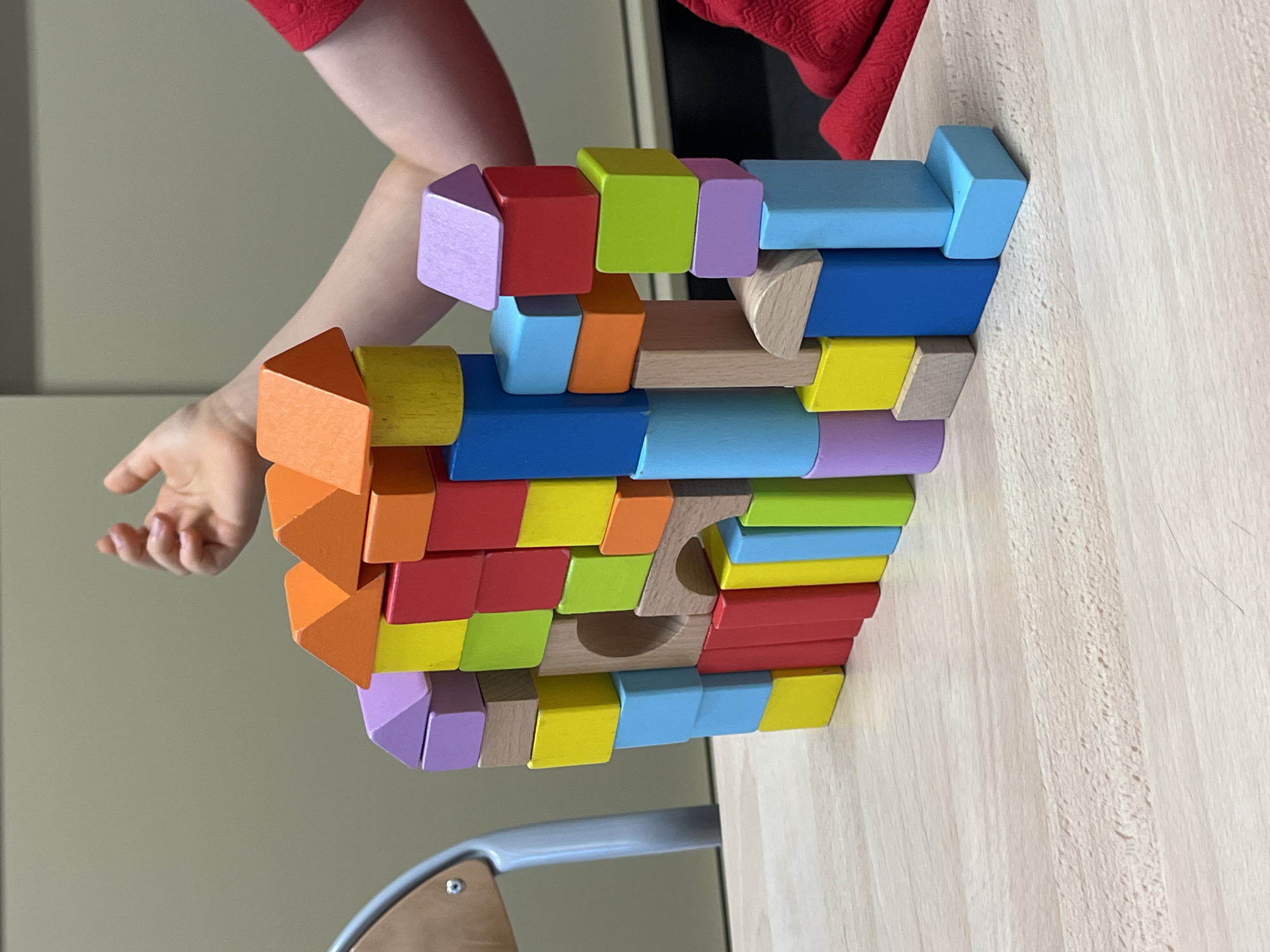The image depicts a colorful tower of wooden blocks constructed on a light-colored wooden table. The blocks are of various shapes and sizes, including rectangular prisms, cubes, and triangular prisms, and are painted in vibrant colors such as red, orange, yellow, green, blue, and purple. A small, circular opening is visible in the center of the block structure. In the background, a person's arm and hand are visible, suggesting they are interacting with the blocks. The person is wearing a red garment. The environment appears to be indoors, with a neutral-colored wall in the background and a part of a chair visible on the left. The image does not contain any elements that would indicate a specific location in the city of La Fouillouse, France.
