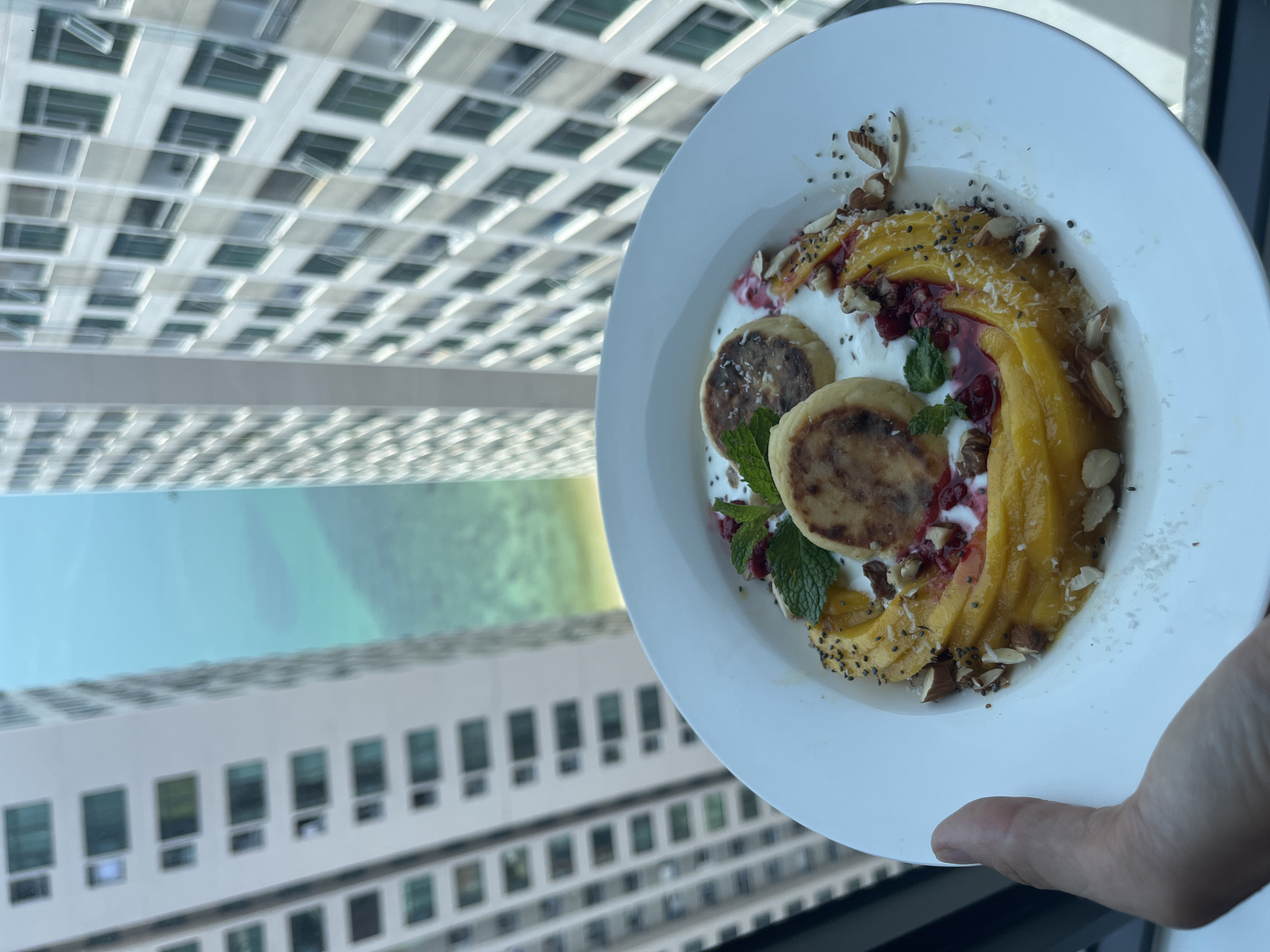 A white plate of food is held in front of a window. The food consists of two small pancake-like items, a dollop of white cream, sliced mango, raspberries, mint leaves, and chopped almonds, sprinkled with chia seeds and coconut flakes. Outside the window, a cityscape includes an ocean view and tall buildings, identifiable as Bac Nha Trang Ward, Vietnam. A person's hand holds the plate. The angle of the shot is slightly askew, as the buildings are tilted to the left.