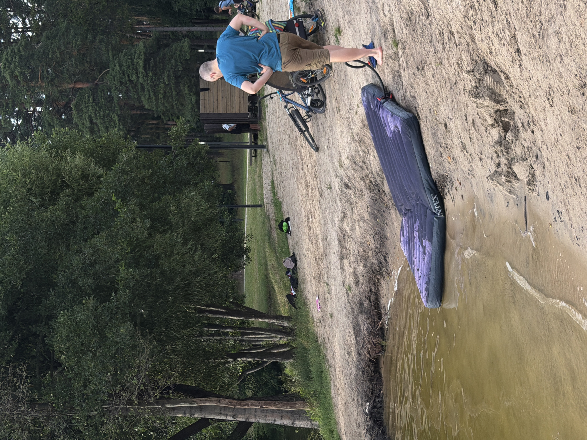 In Malakhovka, Russia, a man is seen on a sandy embankment near trees and a body of water. He is wearing a blue t-shirt and brown shorts, and appears to be using a foot pump connected to an inflatable mattress or sleeping pad. The mattress is dark purple with a subtle pattern. A bicycle is parked nearby. The scene suggests an outdoor recreational activity, possibly camping or preparing for an outdoor adventure by the water. The lighting indicates it is daytime, and the weather appears to be mild. In the background, there are more trees, a small wooden structure, and other people, suggesting a public area like a park or beach. Some personal belongings, including a backpack and a pink item, are scattered on the sand in the middle ground. The "Intex" logo is visible on the inflatable mattress.