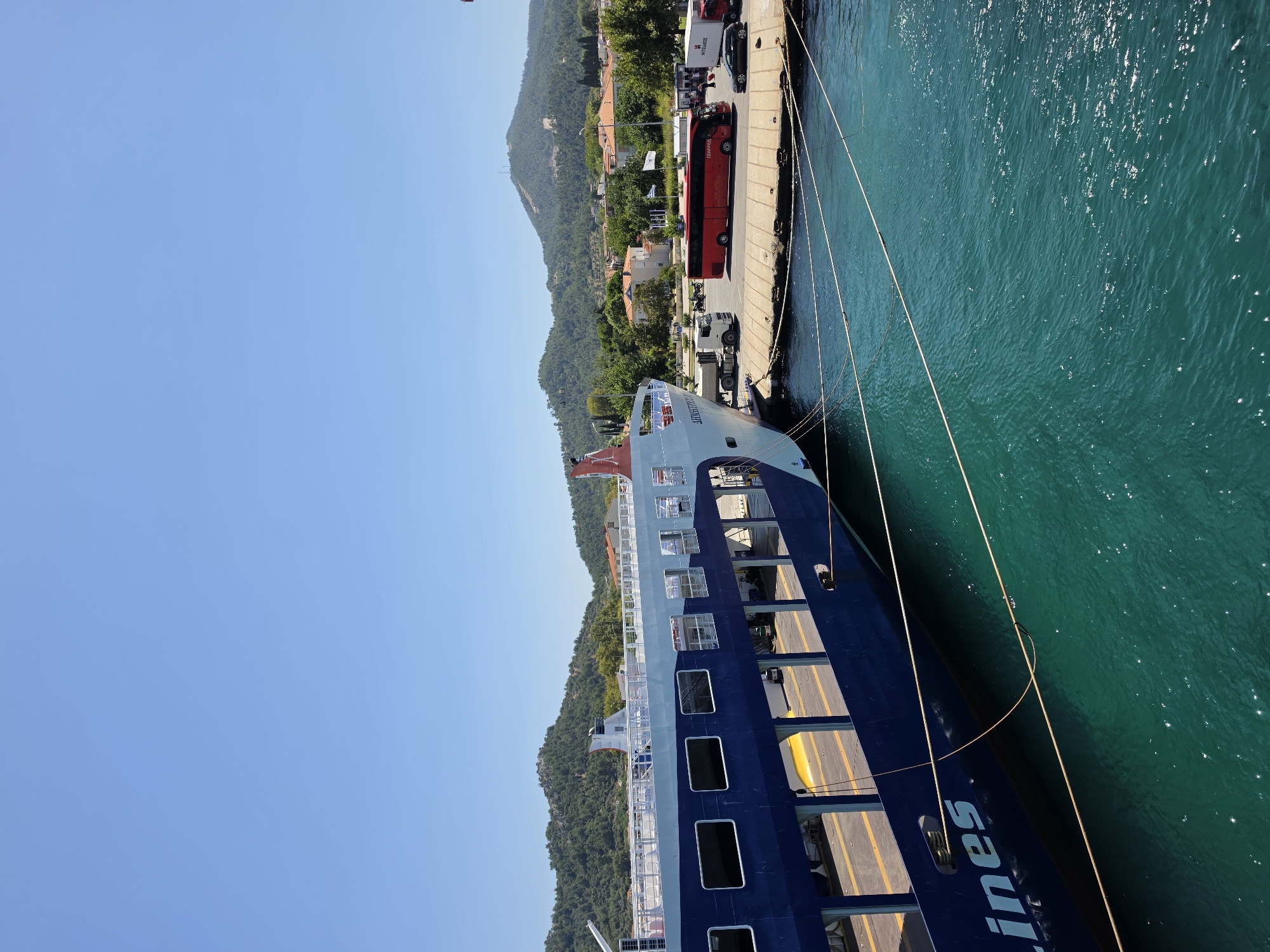 An eye-level, wide shot captures a large blue and white ferry docked at a port under a clear blue sky. The ferry, identified by the partially visible "lines" on its hull, occupies the left side of the frame, with its ramp extended onto the dock. On the dock, a red bus and several trucks are visible, along with a few people near the bus. Behind the dock, a town with red-roofed buildings is nestled at the base of a lush green, hilly landscape. The water in the foreground is a vibrant turquoise.