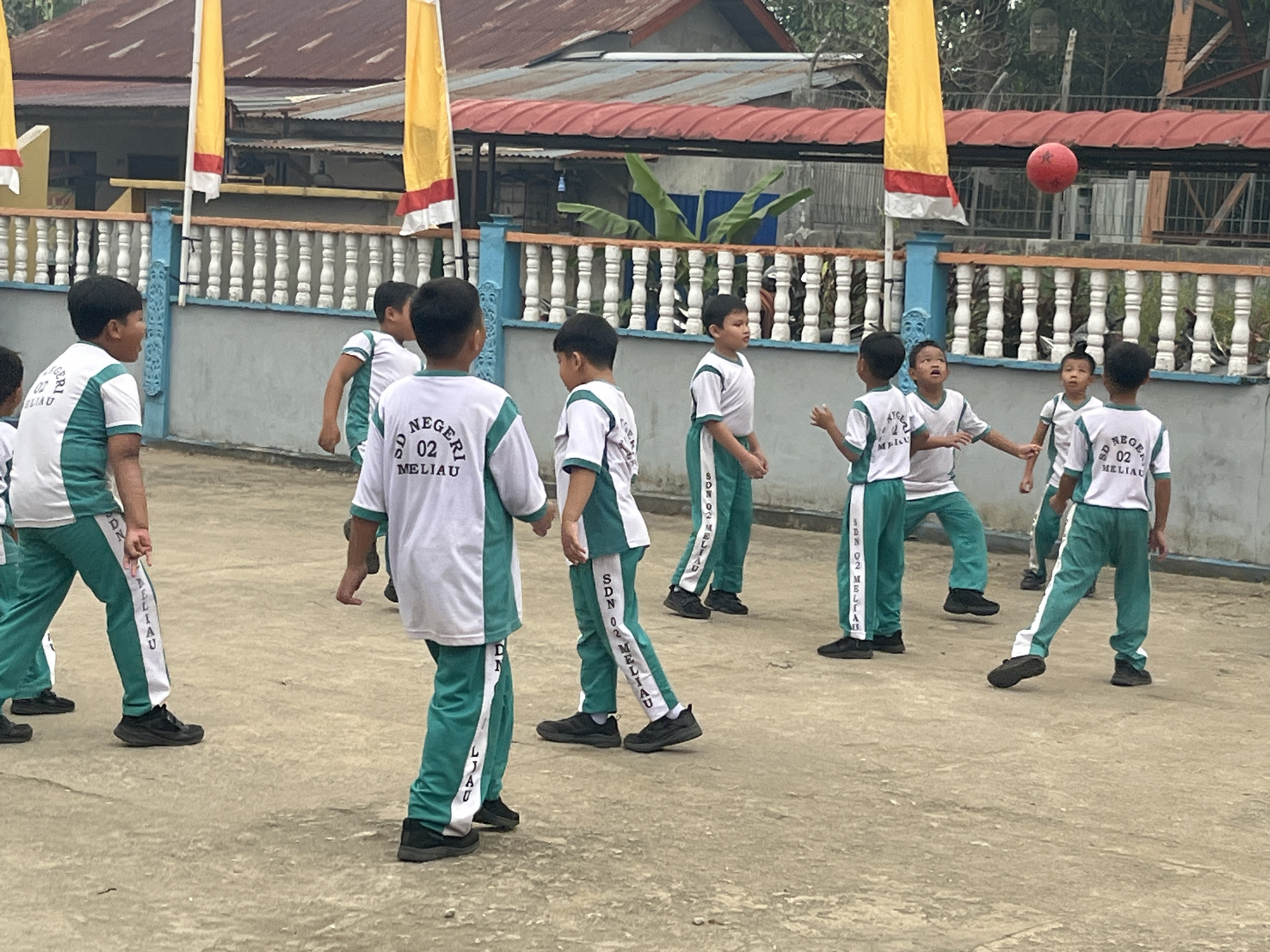 A group of nine young boys, appearing to be elementary school students, are gathered on a paved outdoor area in front of a building with a red tiled roof. They are wearing matching white shirts with green trim and green pants with white stripes. The white shirts have the text "SD NEGERI 02 MELIAU" printed on the back in dark lettering. The green pants also have "SDN 02 MELIAU" printed vertically along the side stripe in white lettering. The boys are actively engaged in a game, with a red ball suspended in the air above some of them, suggesting they are playing some form of catch or a ball game.
The environment includes a white picket fence with a blue railing in the background, and two large yellow flags with red and white trim are visible hanging from poles. Behind the fence is a building with a corrugated red roof, and some greenery is visible. The ground is a light brown concrete surface. The overall impression is a schoolyard scene in Meliau Hulu, Indonesia, where children are participating in outdoor recreational activity.