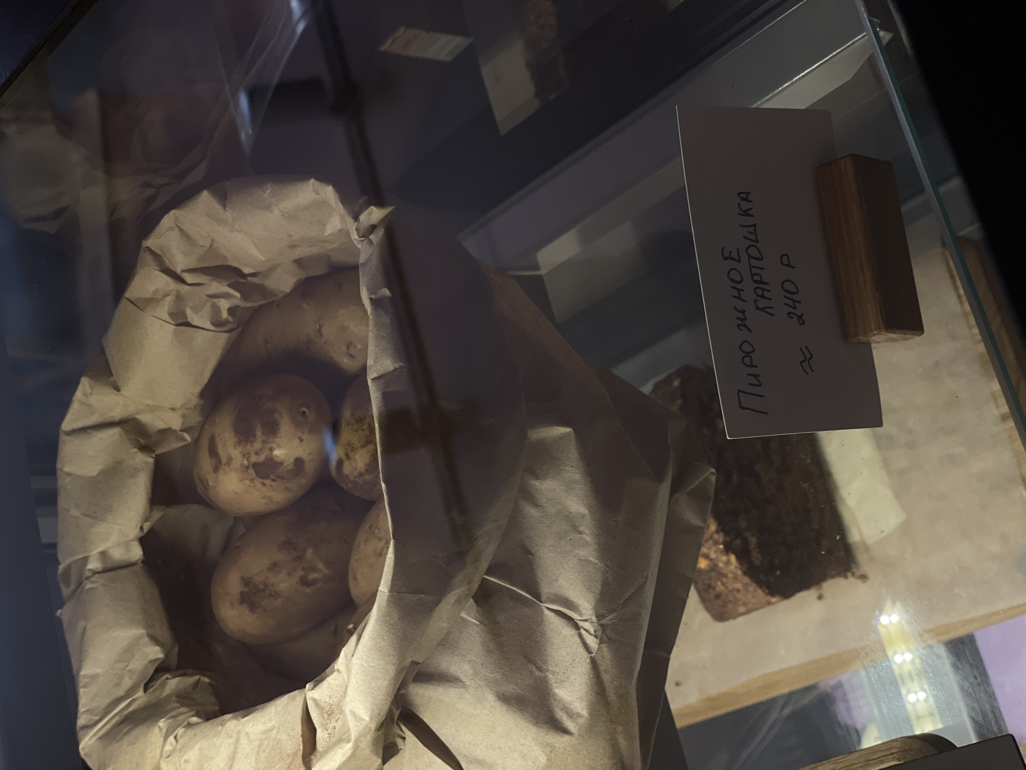 The image shows a display case containing potatoes in a brown paper bag next to a sign that reads "Пирожное картошка ≈ 240 P". This translates to "Potato cake ≈ 240 rubles". The location is likely Krasnoyarsk, Russia.