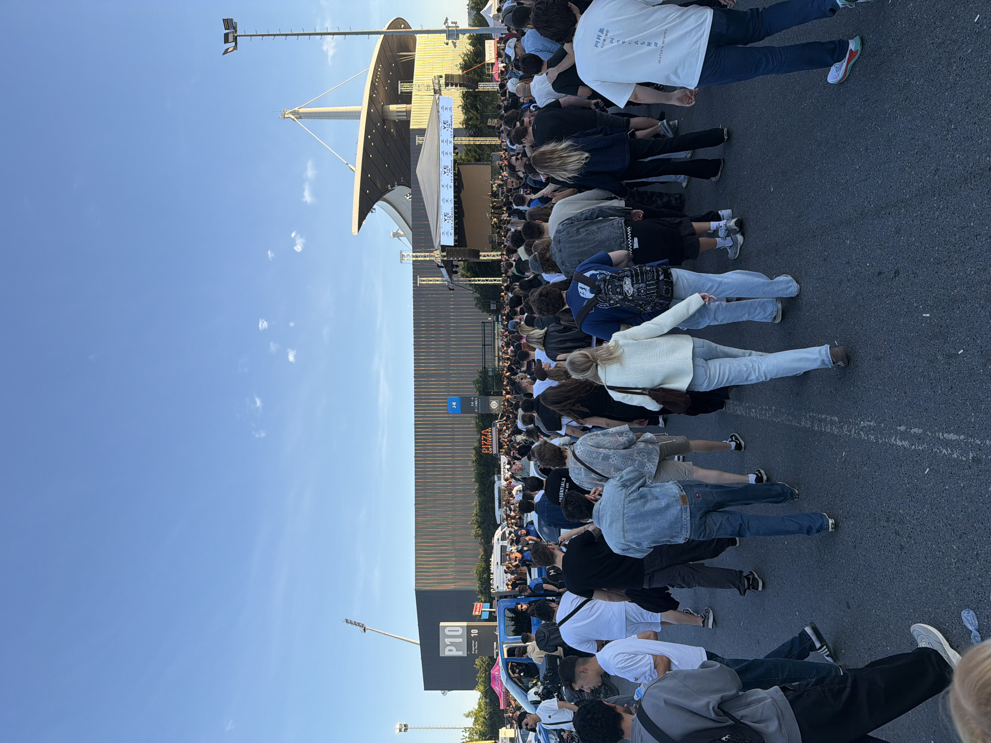 A large crowd of people are gathered in an outdoor area in Başakşehir, Turkey. They are positioned in front of a stage, suggesting they are attending an event or concert. Behind the crowd is a multi-story structure with a unique, curved architectural design. In the lower left, there's a parking sign labeled "P10." The sky is clear and blue. The area appears to be paved and open.