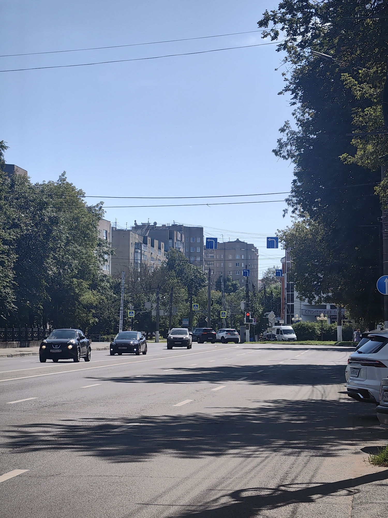 The image depicts a sunny day on a multi-lane road in the city of Tver, Russia. The road is paved and marked with white lane lines. Several vehicles are visible, including black sedans, a black SUV, a white van, and a white SUV. Some vehicles are in motion, while others appear to be parked or stopped. Pedestrian crossing signs are present on the side of the road, and a traffic light is visible. Trees line the left side of the road, providing shade in some areas. Apartment buildings and other structures are visible in the background. The sky is clear and blue.