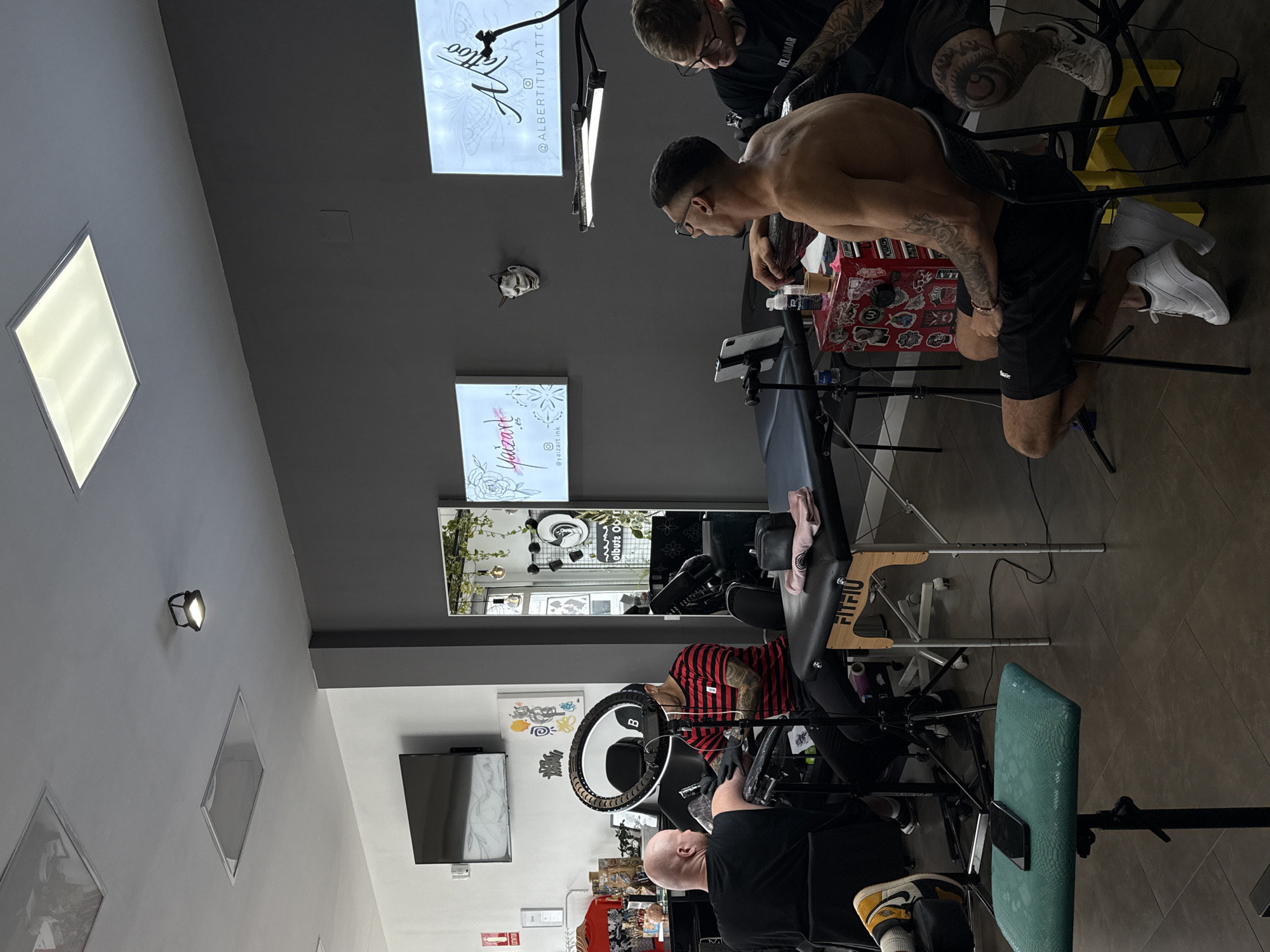 The photograph shows an interior view of what appears to be a tattoo studio. Two tattoo artists are actively working on clients. The studio has gray and white walls.
One artist, wearing a black shirt, is working on a client in a striped shirt seated in a chair illuminated by a ring light. Another artist, also in black, is working on a shirtless client who is leaning forward. Light boxes with tattoo shop logos are on the wall. Other objects include mirrors, tattoo machines, and various tattoo-related supplies.
The presence of a light box with "yaizart.ink" suggests this is a business located in Yaiza, Spain.