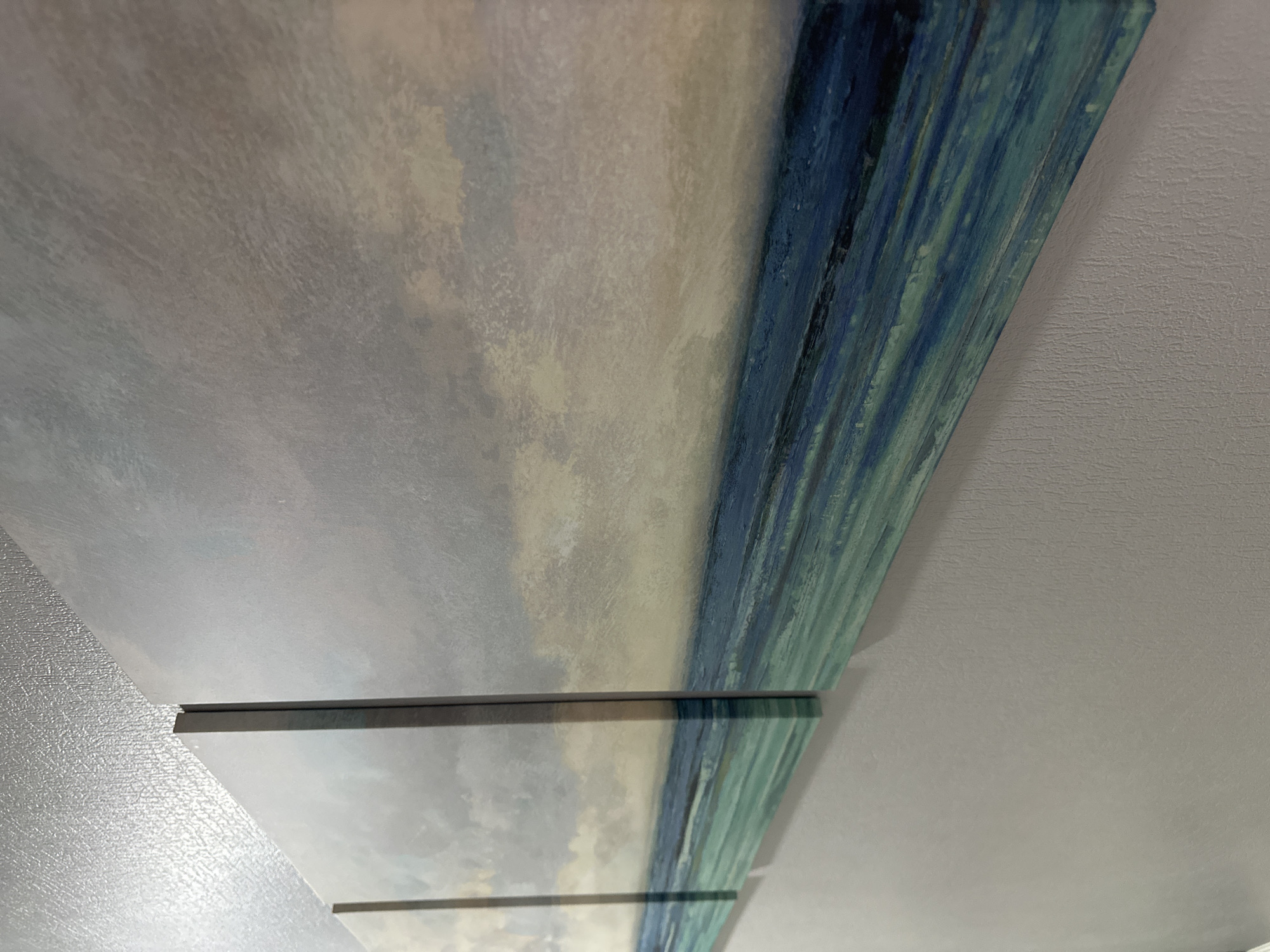 The image shows a close-up, angled view of a triptych painting depicting a seascape. The painting is mounted on a light gray wall, and the textured, silvery ceiling is visible above it. The artwork features abstract brushstrokes of blue, teal, and green in the water, with a hazy, light-colored sky. The three panels are distinct and appear to be spaced slightly apart. The scene is indoors, and there are no people or other objects visible besides the artwork and the wall and ceiling. The location of Nevinnomyssk, Russia, is not depicted or suggested by any elements within the image.