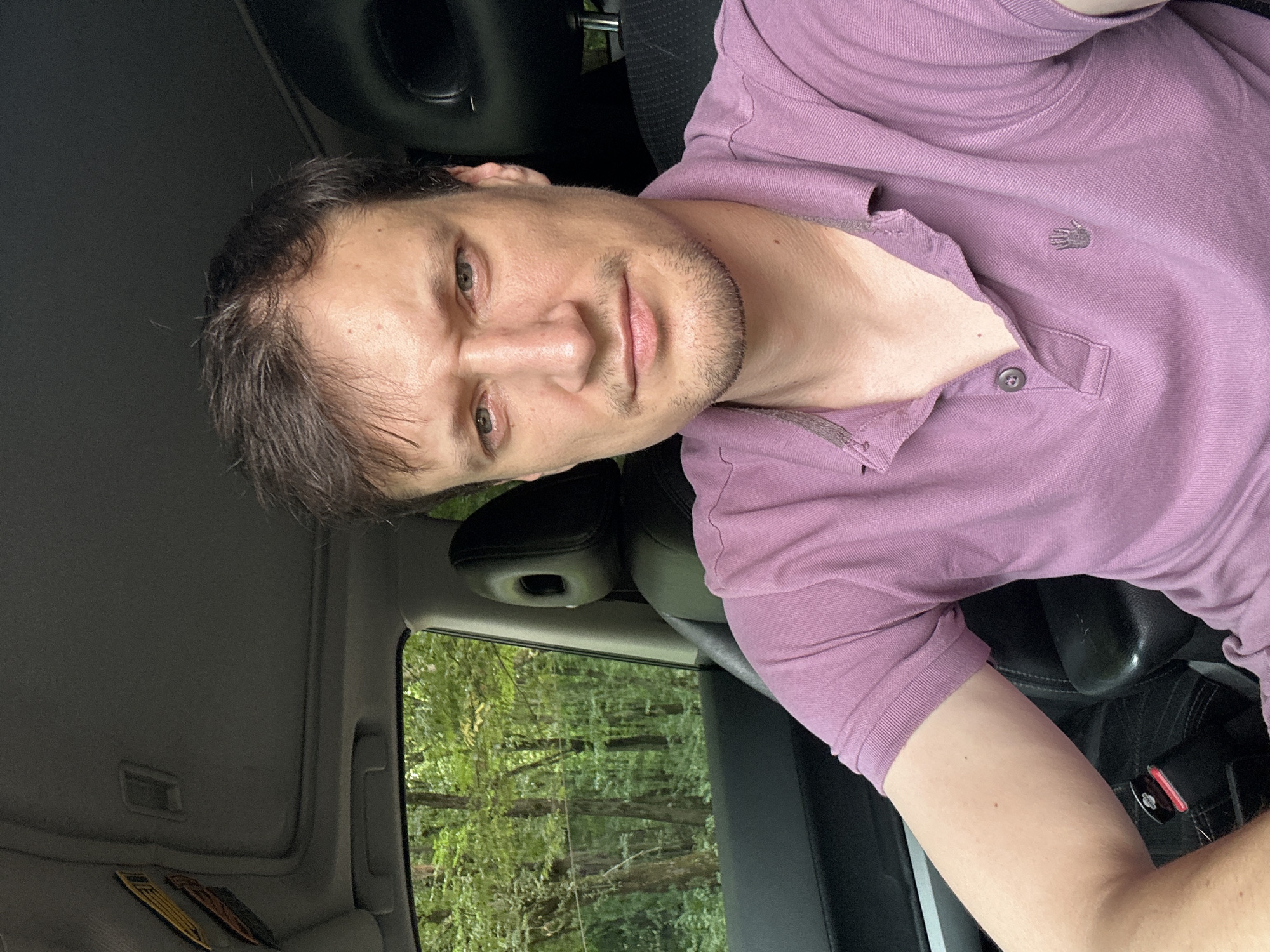 The image shows a man sitting in the driver's seat of a car, with the car interior visible. He is wearing a purple polo shirt. Through the car window, a forest with green trees is visible. The man's face is visible, and he has short, dark hair and a light complexion. The car appears to be parked on a road or path surrounded by trees. The image does not contain any information that would indicate the location is in the city of Shatava, Ukraine.