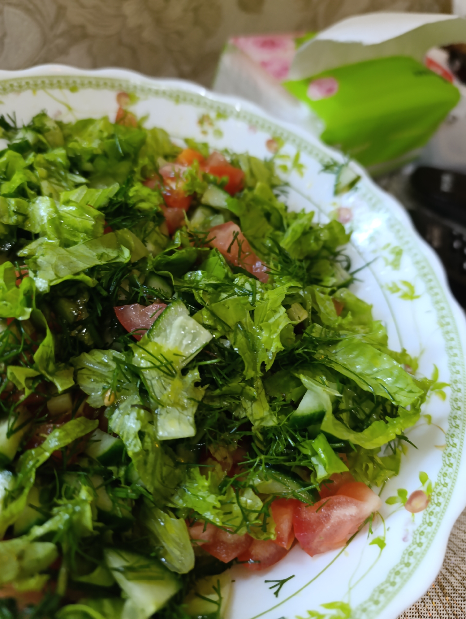 A close-up, slightly high-angle shot captures a serving of fresh salad in a white, oval bowl with a decorative green and floral rim. The salad consists of chopped lettuce, diced tomatoes, and sliced cucumbers, interspersed with finely chopped dill. The ingredients appear moist, suggesting they may have been dressed. In the background, slightly out of focus, is a partially opened white paper bag with a green interior and pink floral patterns. The bag is positioned to the right of the bowl, and beyond it, the environment appears to be an indoor setting with a textured, possibly fabric, surface underneath the bowl. The location of Tugulym, Russia, is not visually evident in the provided image.