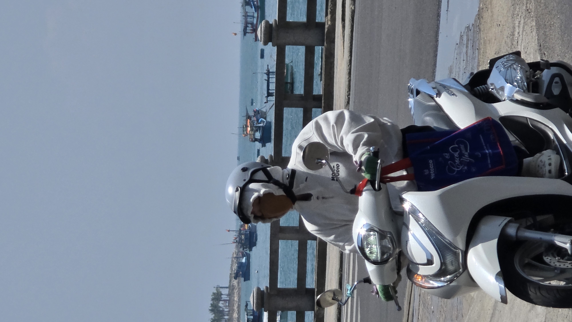 A medium, vertical shot shows a person on a white scooter on a bridge next to an ocean with fishing boats.
The person is wearing a white sweater, helmet, and gloves. The person appears to be looking at the ocean. A blue tote bag sits on the scooter between the person and the handlebars. The ocean has fishing boats in the distance. A bridge or pier rail is to the left of the person. The sky is pale blue. The bridge or pier surface is gray. The image was taken in Bắc Nha Trang, Vietnam.