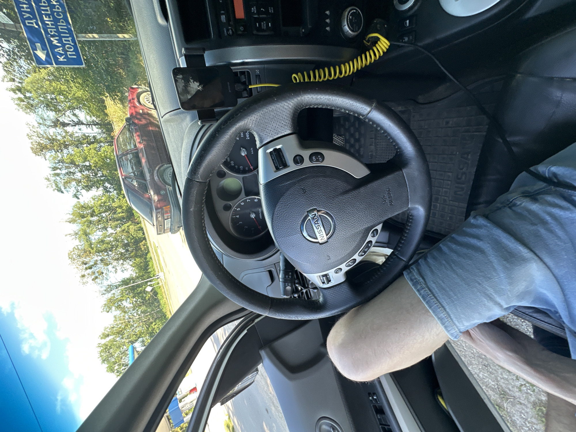 The image is taken from the interior of a Nissan vehicle, looking forward and slightly to the right. A person's leg, clad in light blue denim shorts, is visible in the lower left portion of the frame. The driver's side door panel and a portion of the steering wheel are also visible. The steering wheel has a Nissan logo in the center and control buttons on the left side. A smartphone is mounted on a holder attached to the dashboard to the right of the steering wheel, with a yellow coiled cable extending from it. The dashboard itself is dark.
Outside the car, visible through the windshield, is a road and a red car parked on the side. There is a blue and white road sign pointing left, with text that appears to be in Ukrainian. The text reads "ДУНА" at the top and "КАМ'ЯНЕЦЬ-ПОДІЛЬСЬК" below it, indicating directions to Dunayivtsi and Kamianets-Podilskyi, cities in Ukraine. The location is identified as Shatava, Ukraine. Trees and vegetation line the roadside, and the sky is bright with some clouds.