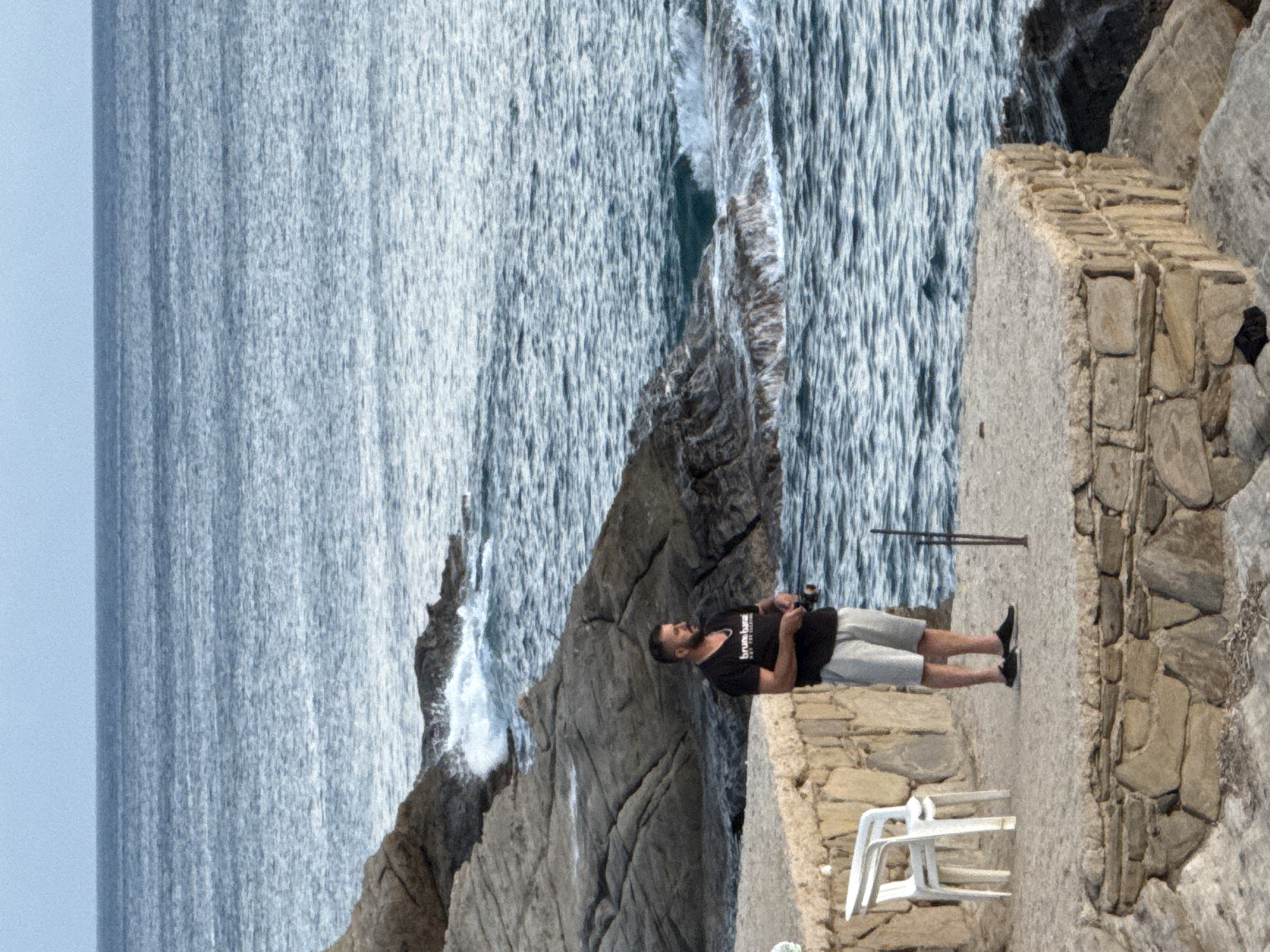 A man stands on a stone-walled promenade overlooking the Aegean Sea in Menderes, Turkey. He is holding a fishing rod and appears to be fishing. The man is wearing a black t-shirt and light-colored shorts. To his left, a white plastic chair is placed on the promenade. The sea is choppy with waves breaking against the rocky shoreline. The sky is overcast.