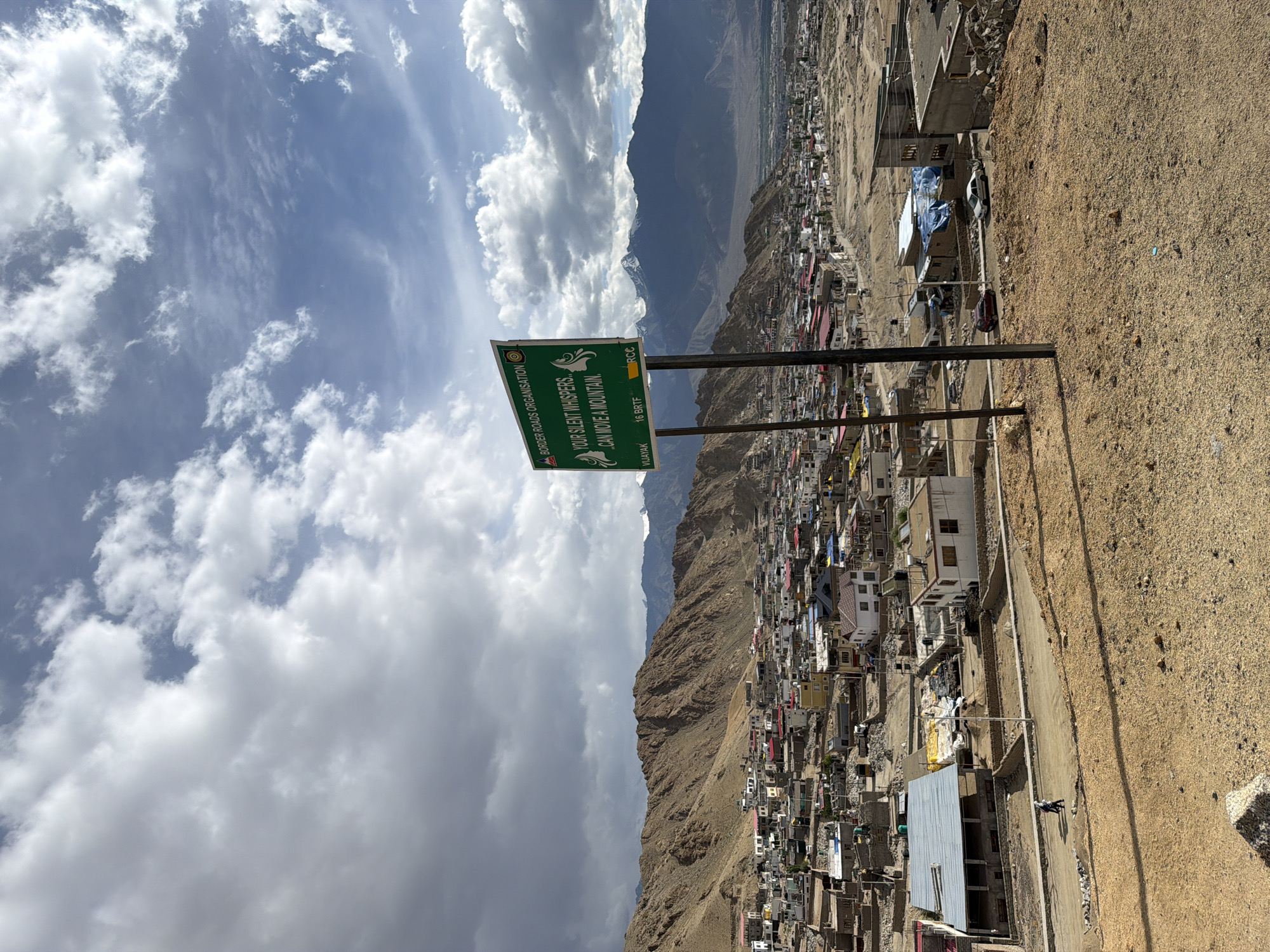 The image shows a panoramic view of the city of Leh, India, nestled in a valley surrounded by brown, arid mountains. The sky is partly cloudy with dramatic formations of white and grey clouds against a blue sky. In the foreground, there is a dry, sandy ground with scattered rocks.
A prominent green signpost stands in the mid-right of the image. The sign reads "BORDER ROADS ORGANISATION" at the top, followed by the text "YOUR SILENT WHISPERS CAN MOVE A MOUNTAIN." There are also smaller inscriptions at the bottom of the sign: "VIJAYAK", "16 BRTF", and "RCC". The sign is mounted on a metal pole.
The city of Leh stretches across the valley floor, characterized by a dense cluster of buildings with flat roofs, many of them painted in white, red, and yellow. Some buildings appear to be constructed from stone and mud. Roads wind through the urban landscape. In the background, larger, snow-capped mountains rise majestically, indicating a high-altitude location.
Several vehicles, including cars and what appears to be a truck or bus, are visible on the roads within the city. Two individuals are walking on a dirt road in the foreground, near a building with a corrugated metal roof. The overall scene depicts a rugged, mountainous environment with a built-up settlement.
