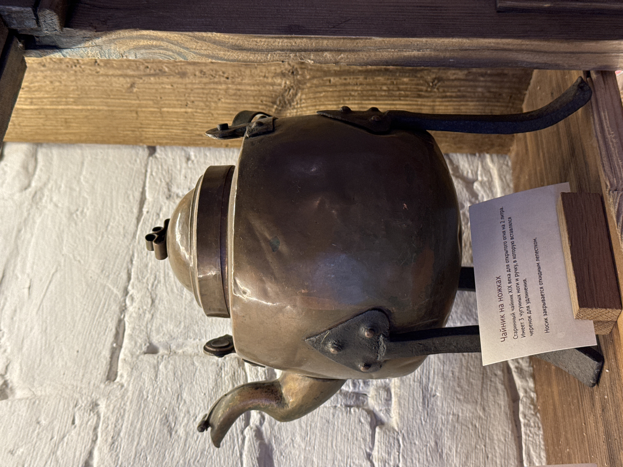 The image shows an antique teapot with a spout, legs, and a handle resting on a wooden ledge. A label next to the teapot reads in Russian about the teapot being from the 19th century, used for open fire, with a capacity of 2 liters. The text also describes the cast iron legs and handle, with a wooden extension that can be inserted. The background includes a rough white brick wall and wooden beams. It appears to be located in the city of Novo-Nikol'skoe, Russia.