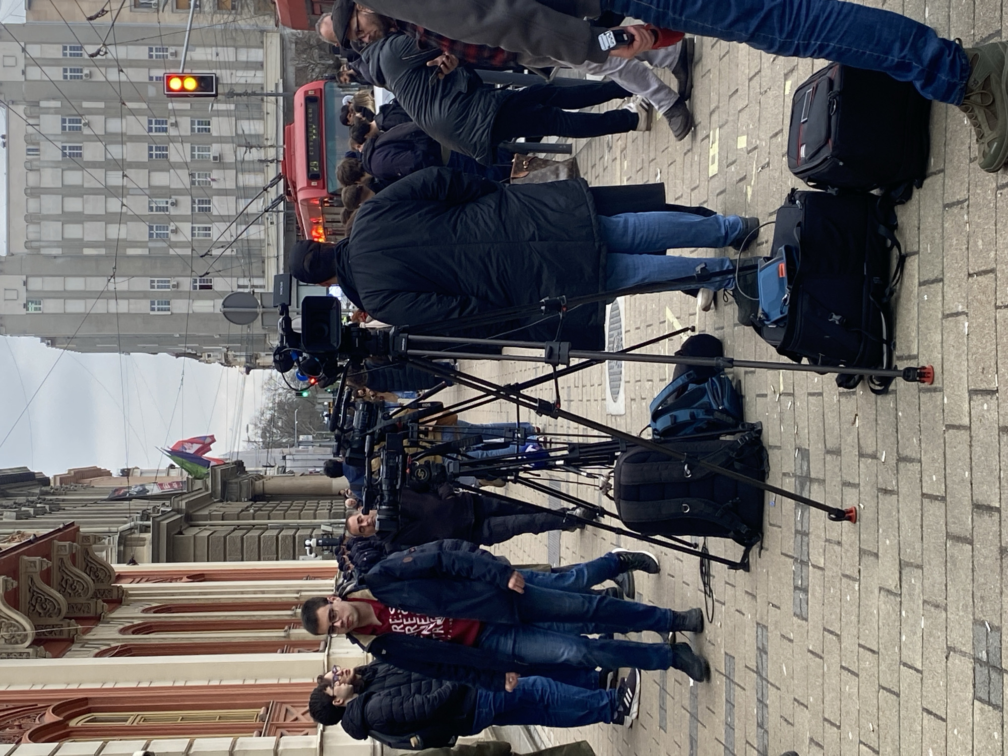 A group of people, including journalists with professional video cameras on tripods, are gathered on a sidewalk in Belgrade, Serbia. In the background, a red tram and a gray building with a traffic light showing a red signal can be seen. Flags are flying on top of a building to the left. People are standing and waiting; the journalists are pointing their cameras in the same direction. Several backpacks and camera bags are on the ground behind the cameras. The sidewalk is made of rectangular gray paving stones. The weather appears to be overcast.