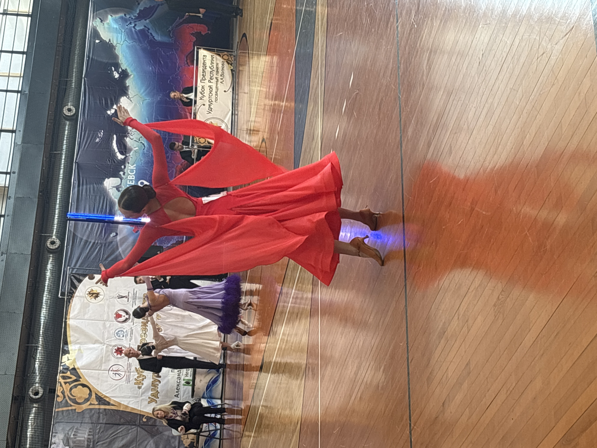 The image captures a ballroom dancing competition, likely in Izhevsk, Russia. The dancers are performing on a polished wooden floor. A woman in a flowing red dress is the focal point, with arms outstretched. Other dancers are visible in the background wearing ballroom gowns. A banner on the wall reads "President of Udmurt Republic Cup in memory of A.A. Volkov." Spectators sit in chairs watching the performance. The event appears to be taking place indoors.
