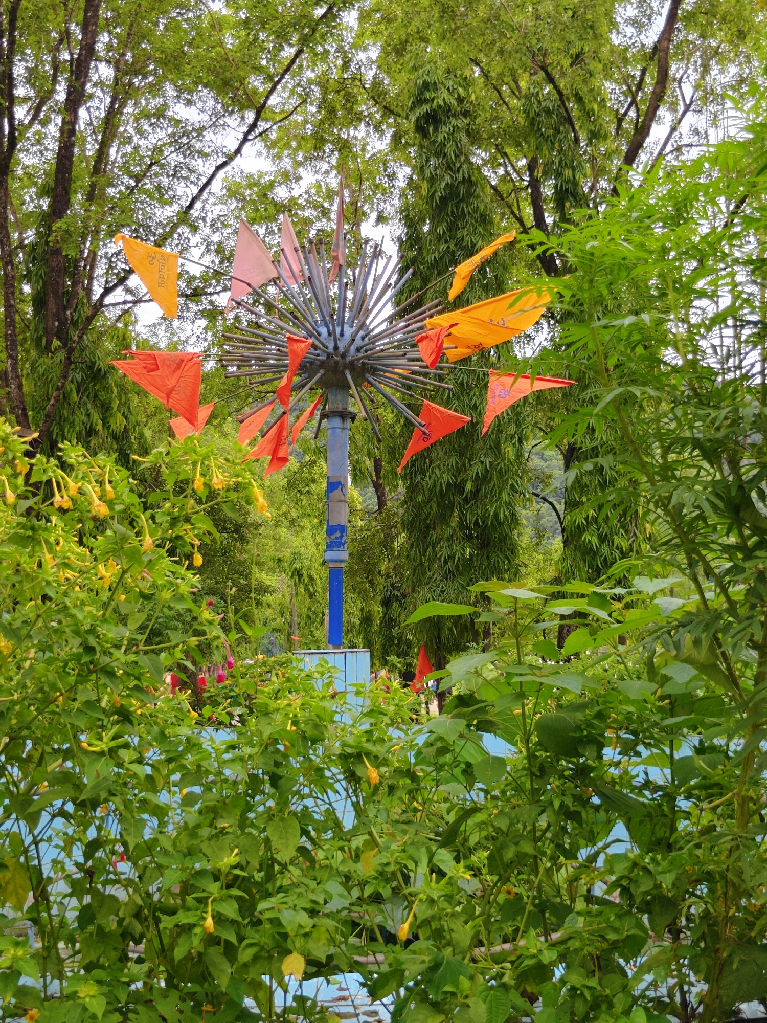 The image shows a vibrant outdoor scene, possibly within a park or garden in Pokhara, Nepal. In the foreground, lush green foliage with small yellow flowers partially obscures the view. Behind the greenery, a metal structure with radiating metal rods is visible, adorned with several triangular flags in shades of orange and pink. A blue pole supports the central structure. In the background, tall trees with dense green leaves form a canopy, creating a natural backdrop. Hints of blue structures and possibly water are visible beneath the foliage and flags, suggesting a recreational area or water feature. The overall impression is one of a colorful and lively outdoor environment, likely a public space within Pokhara.