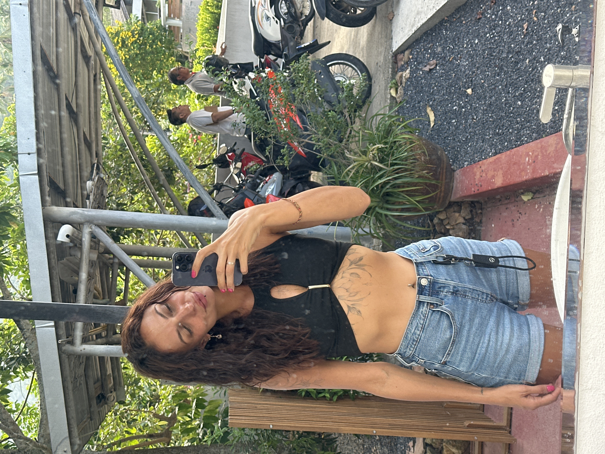 A woman with long, wavy brown hair takes a selfie in a mirror. She is wearing a black crop top with a keyhole cutout and a gold accent, and light-wash denim shorts. Her arms are visible, with a bracelet on her left wrist and a tattoo on her right forearm. Her nails are painted bright pink. She has a piercing on her lip and a tattoo on her abdomen.
The background suggests an outdoor setting, likely a public area. To the left, a wooden structure is visible. Behind the woman, several motorcycles are parked. Two men are standing in the background, one wearing a white t-shirt and the other a grey polo shirt. Lush green foliage and trees are abundant, with some yellow flowers visible on a tree in the middle ground. The ground is covered with dark gravel. A potted plant sits in the foreground. A faucet is visible in the lower right corner, indicating the presence of a sink or water source. The scene appears to be in Ko Samui District, Thailand, suggested by the tropical vegetation and the presence of parked motorcycles, common in the region.