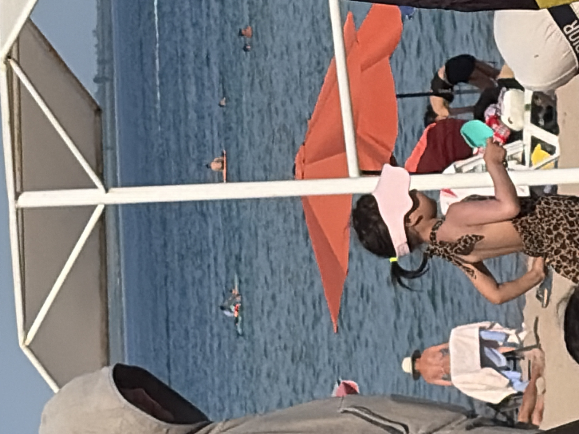 The image shows a group of people on a boat or floating structure on the water. The water appears calm and blue, suggesting good weather. The scene is brightly lit, likely indicating daytime, possibly a sunny afternoon.
In the foreground, a child wearing a pink headband and a leopard print outfit is visible. They appear to be interacting with something out of frame or looking at another person. Next to the child, a red and orange umbrella is partially visible, providing shade.
Further back, other individuals are present. One person is wearing a dark outfit and appears to be bending over. Another individual is wearing a white hat and is partially covered by a white cloth. There are also glimpses of other people scattered across the boat. The overall atmosphere seems relaxed and casual, indicative of a leisure activity such as a day trip or vacation. The setting is on a body of water, with the horizon and a distant shoreline or structure visible in the background. There are no discernible emotions explicitly captured in the image. No text is clearly legible.