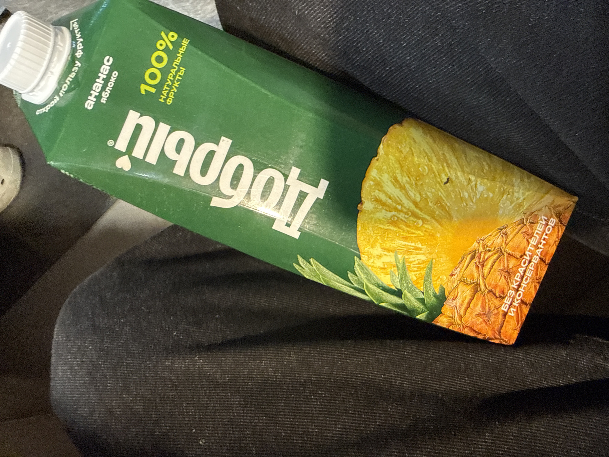 A green juice carton is prominently featured, lying at an angle on a dark, textured fabric surface, possibly clothing or upholstery. The lighting appears dim, suggesting an indoor setting or a low-light environment. The carton is branded "ПЛОДОГОСТИ" (PLODOGOSTI), with the name written upside down in white Cyrillic letters.
The juice flavor is indicated as "АНАНАС ЯБЛОКО" (ANANAS YABLOKO), meaning Pineapple Apple. A bright yellow slice of pineapple and a whole pineapple with green leaves are depicted on the front of the carton. Further text states "100% НАТУРАЛЬНЫЕ ФРУКТЫ" (100% NATURALNYE FRUKTY), which translates to "100% Natural Fruits." Another claim, "БЕЗ КРАСИТЕЛЕЙ И КОНСЕРВАНТОВ" (BEZ KRASITELEY I KONSERVATOV), specifies "Without Dyes and Preservatives." A small phrase near the white screw cap reads "открой пользу фруктов" (otkroy polzu fruktov), meaning "discover the benefit of fruits."
No people are visible. The carton appears to be casually placed, perhaps being transported or temporarily set down. The scene is static, without any active event taking place. The presence of Cyrillic text and the location context of Birobidzhan, Russia, confirm the product's origin and potential consumption area.