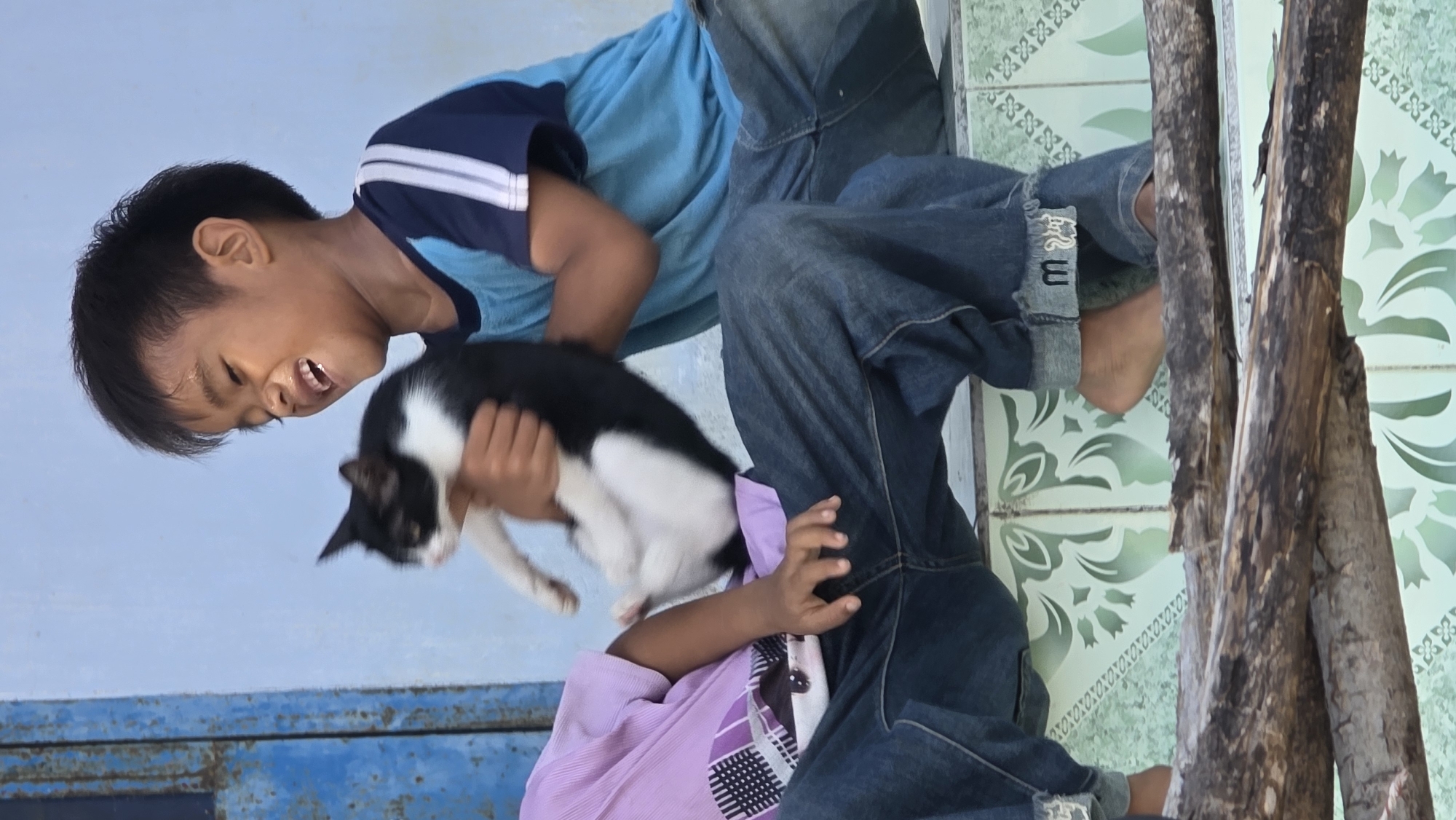 Two children are holding a black and white cat outdoors in Ba Ngoi Ward, Vietnam. One child wears a blue shirt, while the other wears a light purple shirt and blue jeans. The background includes a blue wall and patterned tile flooring. A stacked wood pile is visible on the right.