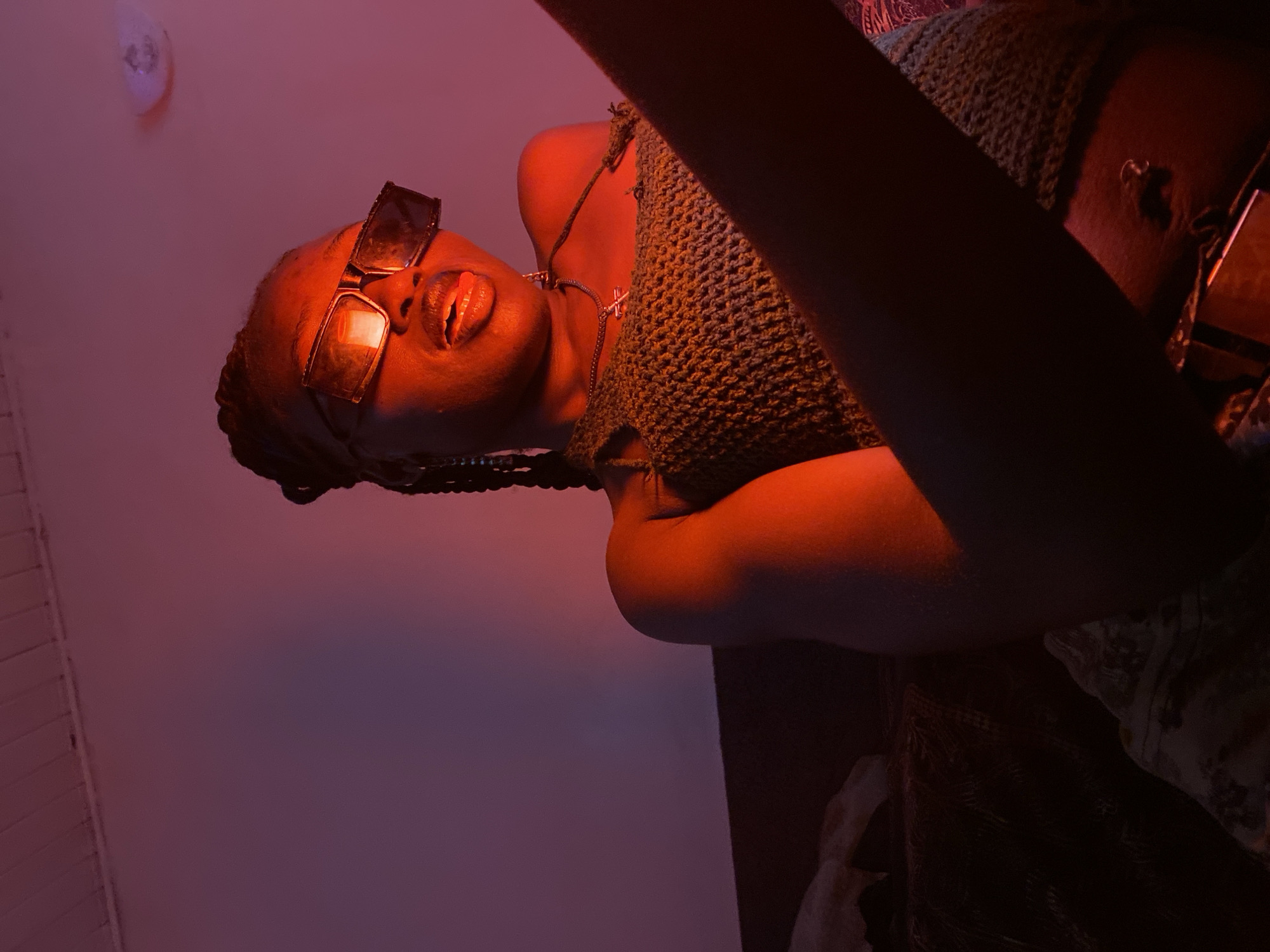 The image shows a young Black woman with braided hair, wearing large, dark sunglasses and a crocheted halter top. She is indoors, illuminated by red and purple lighting, which casts a warm glow on her skin and the background wall. The woman is seated, with her arm extending towards the camera, and she appears to be looking slightly off-camera. The environment is dimly lit, with hints of furniture and patterned fabric visible in the foreground and background. The image is a portrait taken from a medium close-up perspective. The location of Bar Jirgi Summa, Nigeria, is not determinable from the visual information provided in the image.