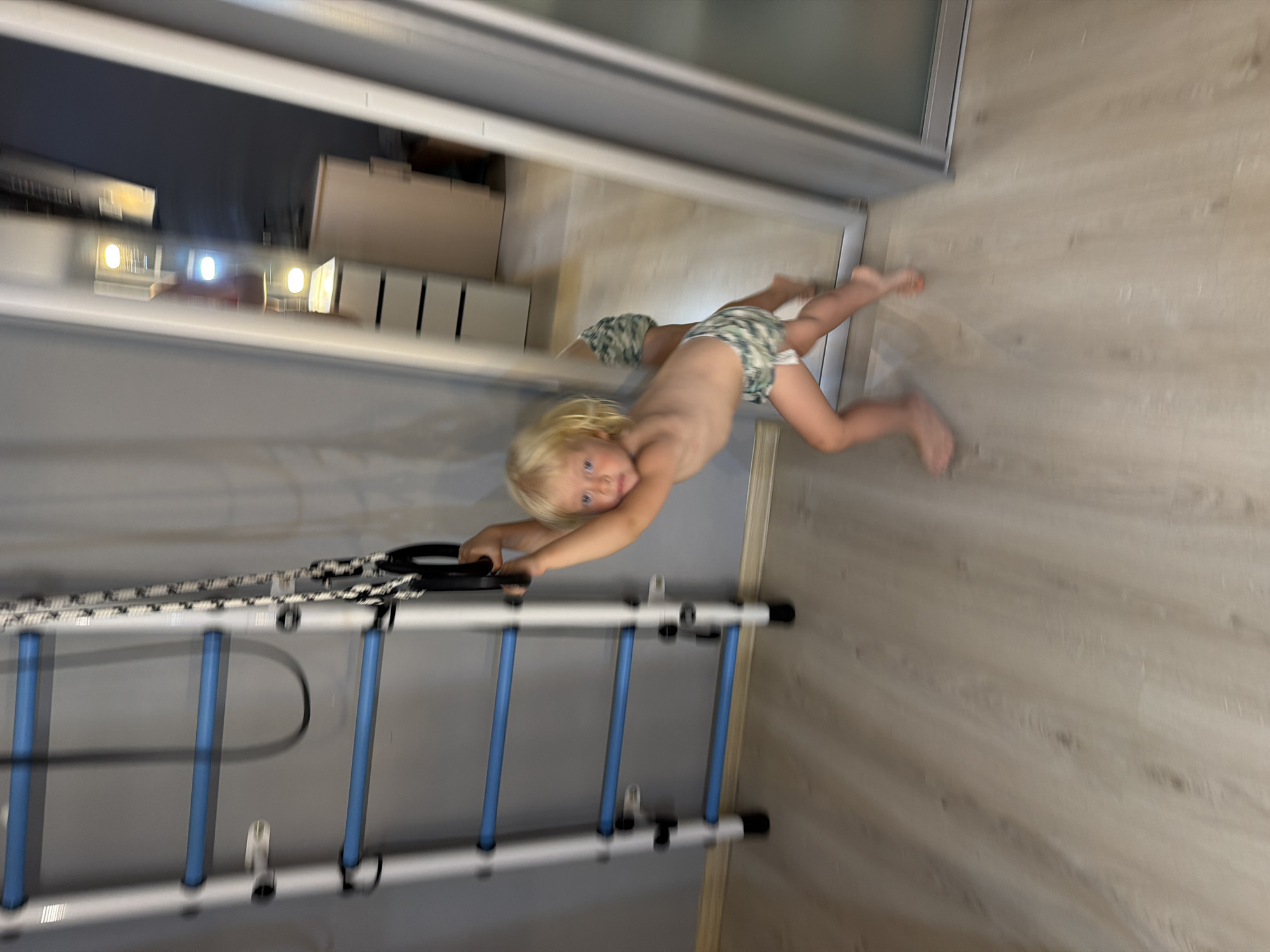The image depicts a young child with blonde hair, wearing camouflage shorts, hanging from gymnastic rings attached to a wall-mounted climbing frame. The child is in motion, creating a blur effect. The climbing frame has white and blue bars and is mounted on a grey wall. The floor is light-colored laminate wood. To the right of the climbing frame, a large mirror reflects the scene, showing another view of the child and a dresser with drawers. The overall environment appears to be an indoor room, possibly a home gym or a child's play area. The image was taken in Sochi, Russia.