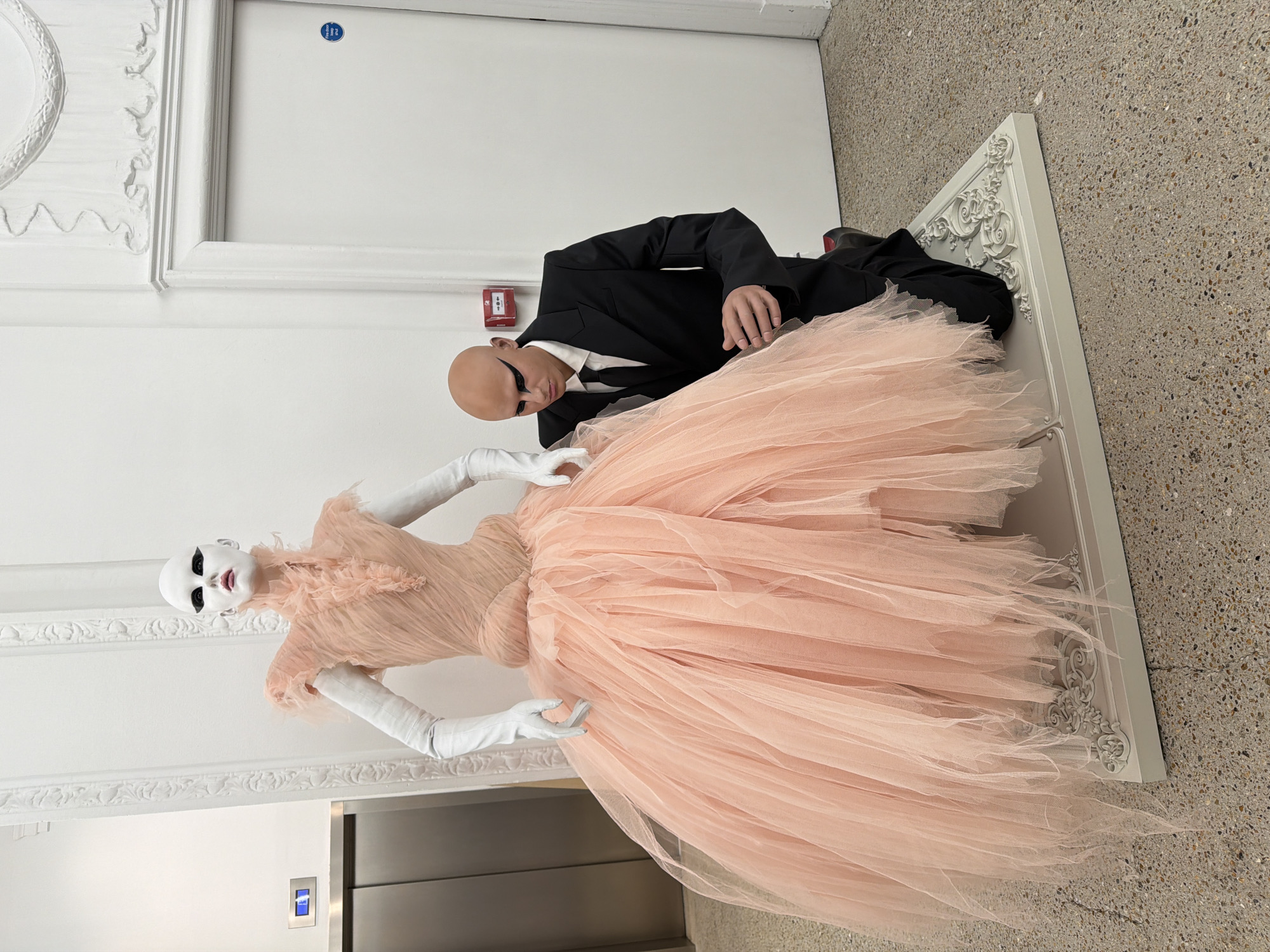 The image depicts a bald man in a black suit kneeling next to a mannequin dressed in a peach-colored tulle gown and white gloves. The mannequin has a white painted face and heavy black eyeliner. They are situated in what appears to be a modern, ornate building in London, United Kingdom, characterized by decorative white moldings along the walls and ceiling, and a terrazzo floor. An elevator is visible on the left side of the frame. A white, decorative frame is positioned on the floor near the mannequin. The man is positioned to appear as if he is attending to or admiring the mannequin.