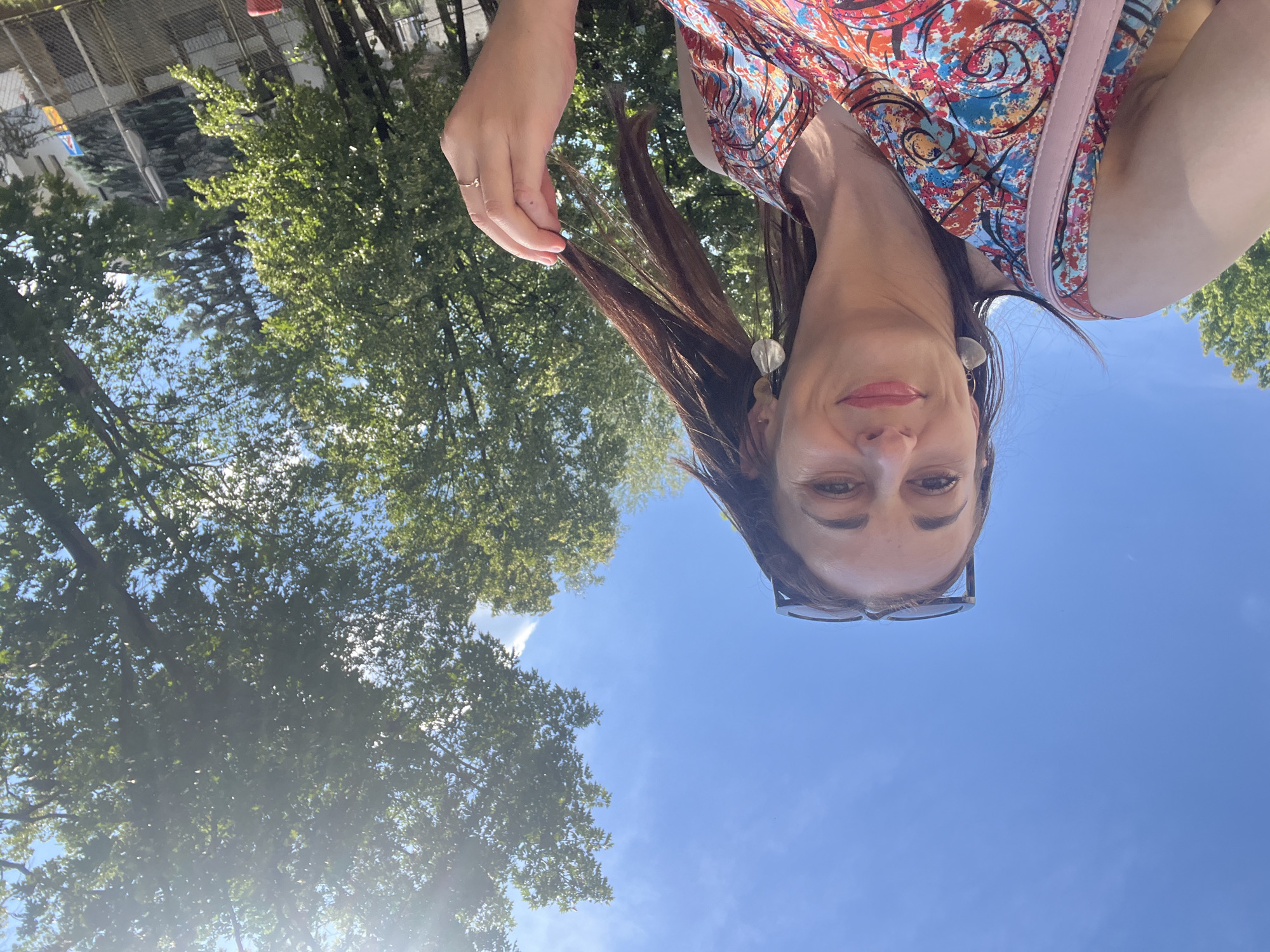 The media file is a selfie taken by a woman outdoors on a sunny day. The subject is a woman with brown hair, wearing a colorful patterned dress and glasses. She is smiling and looking at the camera. Her left hand is raised, touching her hair. In the background, there are green trees and a bright blue sky. The sun is shining, creating some lens flare. The overall impression is one of a casual, happy moment taken during the day. There is no visible text in the image.