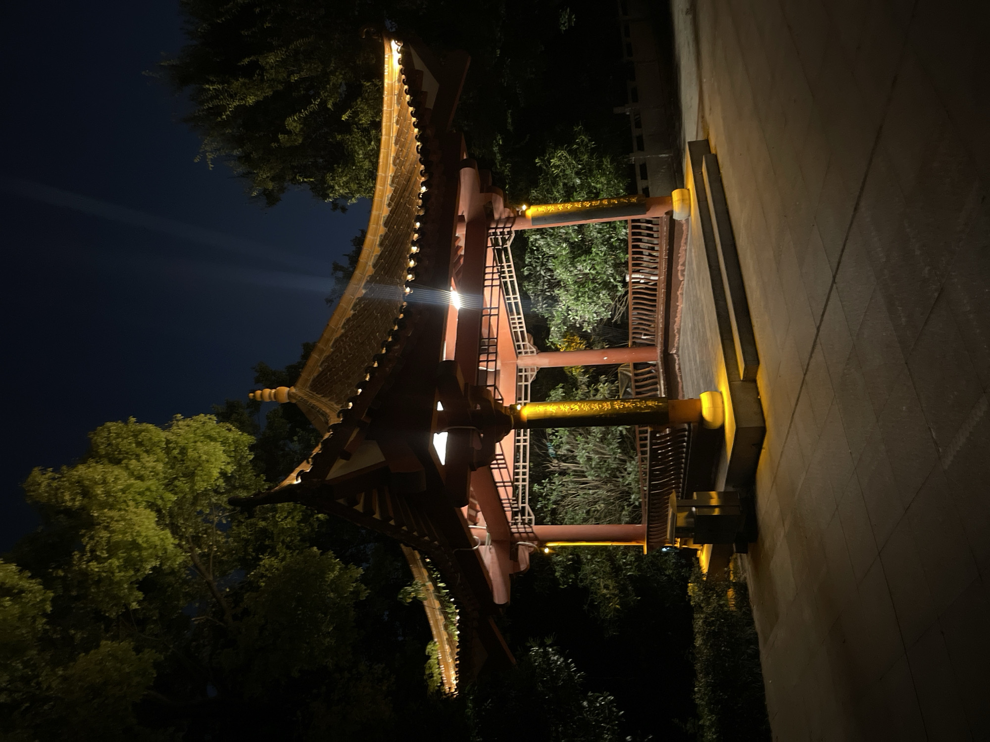 An evening shot captures a traditional East Asian style pavilion, brightly illuminated against a dark night sky. The pavilion, likely constructed of dark red or brown wood, features a gracefully curved, multi-tiered roof with ornate eaves, characteristic of East Asian architecture. Warm, yellow lights line the edges of the roof, glow from within the structure, and accent vertical posts and horizontal beams. Some of these lights softly illuminate the surrounding trees, whose green foliage contrasts with the deep blue-black sky. A faint beam of light is visible cutting across the upper left portion of the sky. On a horizontal beam of the pavilion, golden East Asian script (likely Chinese characters) is visible. The ground to the right of the pavilion is paved with rectangular tiles or stones, also subtly lit by the pavilion's warm glow. The scene is devoid of people, creating a peaceful and serene atmosphere.