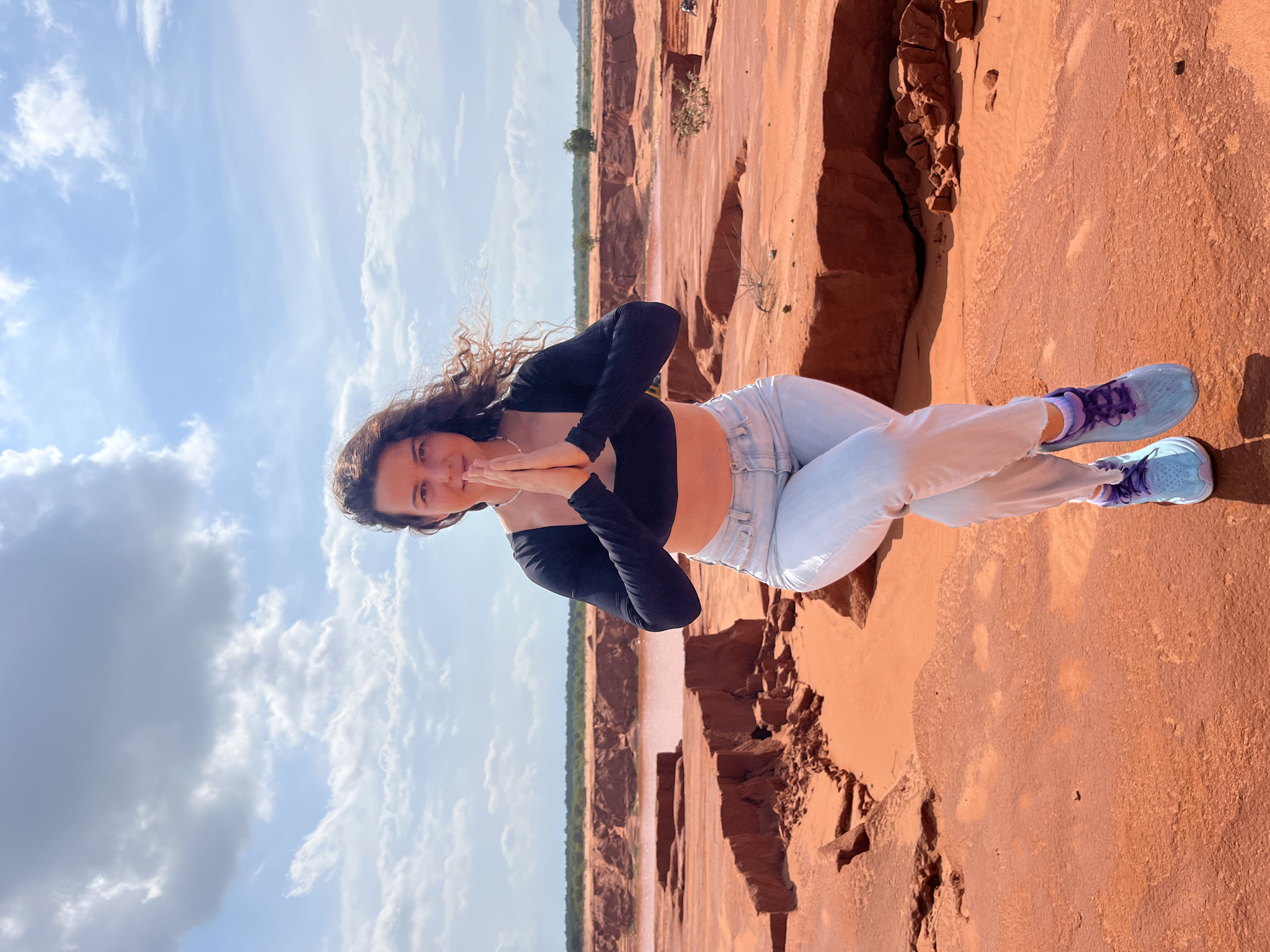 A young woman with dark, curly hair stands outdoors in a desert-like environment with red earth and a body of water in the background. She is wearing a black long-sleeved crop top, light blue jeans, and light blue and purple sneakers. Her hands are clasped in front of her chest in a prayer-like gesture. The sky is blue with scattered clouds. The landscape suggests a location with unique geological formations, possibly eroded by water. The woman is positioned on a ledge of red earth, with more eroded landscape and a body of water visible behind her. The scene is brightly lit by the sun. There is no indication that this scene is located in Mui Ne, Vietnam.