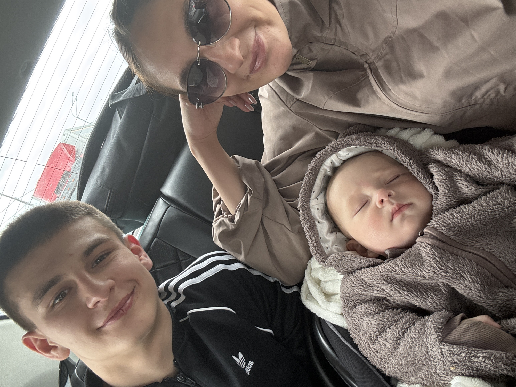 A photo taken inside a vehicle features three individuals: a young man, a woman wearing sunglasses, and a sleeping baby. The young man is seated in the foreground, smiling at the camera. The woman is positioned above the baby, also looking towards the camera. The baby is swaddled in a fluffy, hooded outfit. The interior of the car is visible, with a black leather seat and a glimpse of the vehicle's rear window. Outside the window, there is a building with red and white elements. This photograph was taken in the city of Sochi, Russia.