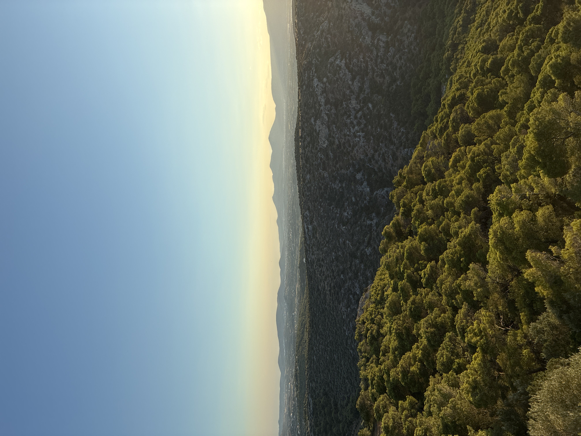 A panoramic landscape view captures the rolling hills and mountains of Limenas Markopoulou, Greece, bathed in the warm glow of late afternoon sun. The sky is a clear, gradient blue, transitioning to a soft yellow near the horizon. In the foreground, a dense forest of green pine trees covers a steep slope, their tops illuminated by sunlight. Beyond the pine-covered hills, a rugged, rocky terrain is visible, also sparsely vegetated. Further in the distance, a hazy expanse of mountains stretches across the horizon, with hints of a distant town or settlement visible between them. The overall scene evokes a sense of natural tranquility and the vastness of the Greek landscape.