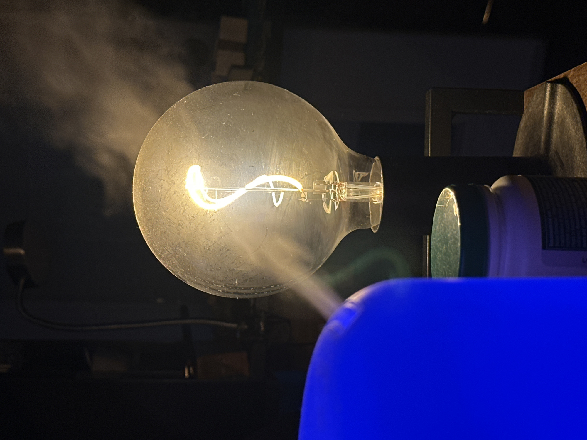 The image captures a close-up of an illuminated incandescent light bulb with a visible filament, emitting a warm, yellow glow. A stream of vapor or mist is seen rising from the right side and dissipating into the dark background. In the foreground, a large, bright blue object, possibly a diffuser or humidifier, dominates the left side of the frame. Partially visible behind it is a white container with a green lid, which appears to be a jar or bottle. The scene is dimly lit, with the light bulb and the blue object serving as primary light sources. The background is dark and indistinct, suggesting an indoor setting, likely a dimly lit room. There are no visible people or animals. The composition is tightly framed, focusing on the interplay of light, mist, and the foreground objects. The location in McMasterville, Canada, is not discernible from the visual elements present in the image.