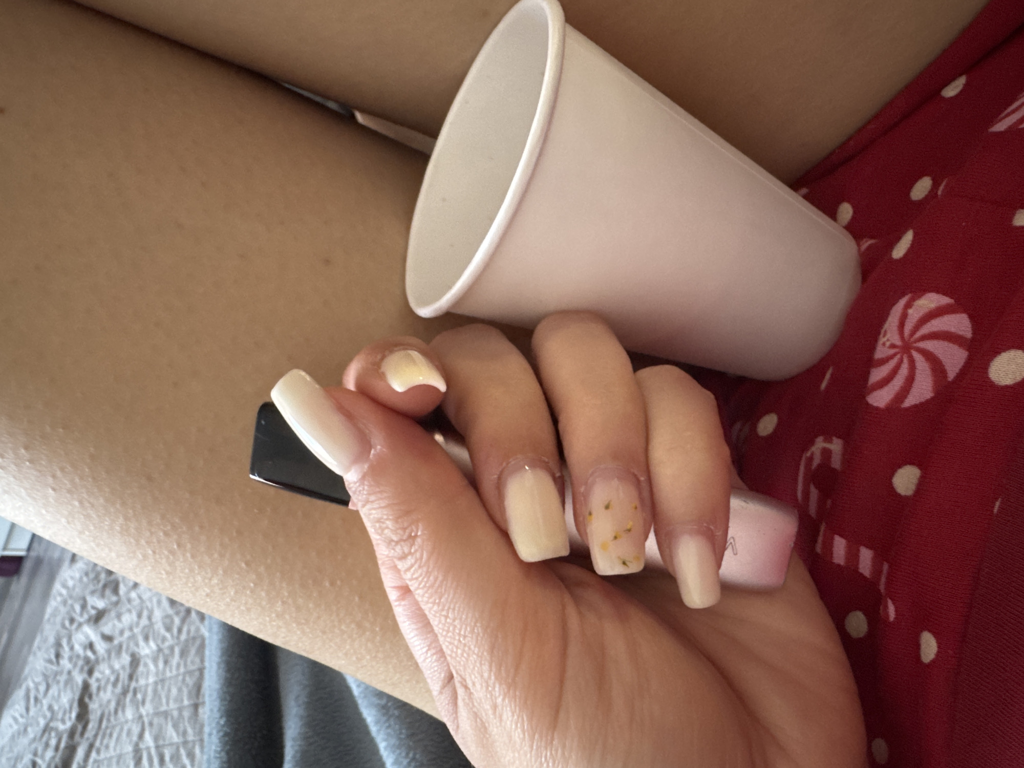 A hand holds a vape pen, with an off-white cup resting on the fingers. The hand is positioned against a background of a person's thigh and a red Christmas-themed fabric.
The location is unknown, and cannot be determined from the image.