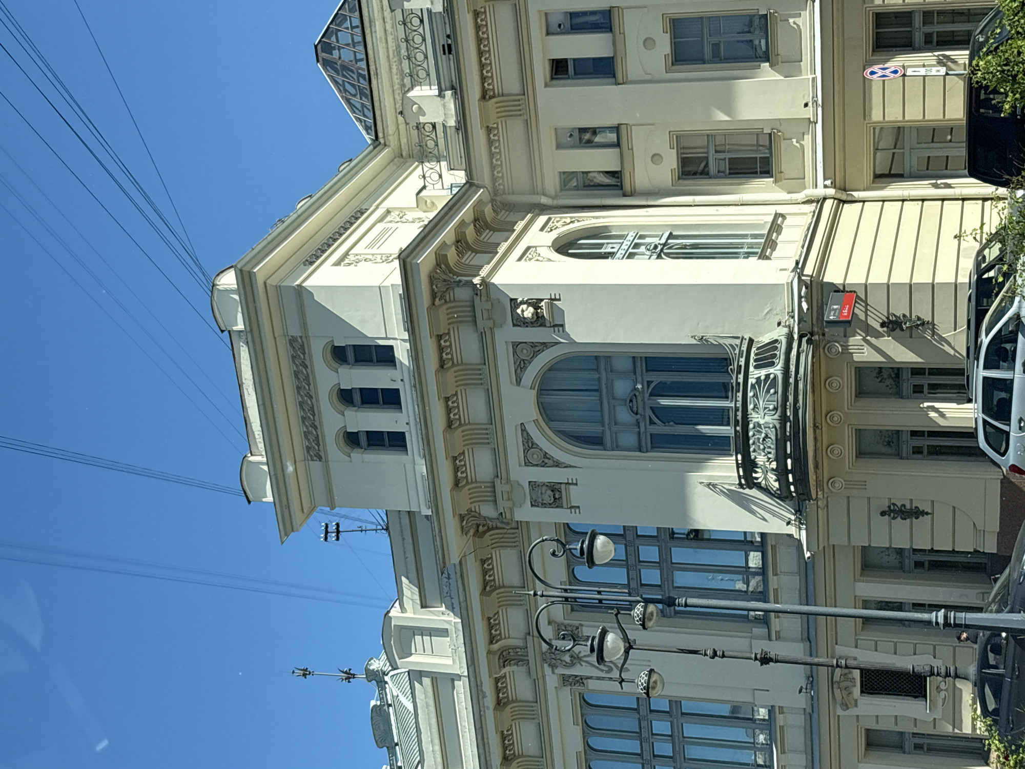 The image depicts a neoclassical building on a sunny day in Saint Petersburg, Russia. The building, painted a light yellow, features elaborate architectural details including decorative moldings, arched windows, and a glass-domed roof section. A distinctive feature is a wrought-iron balcony protruding from the second story. In the foreground, a street lamp with multiple globes stands tall. Several cars are parked along the street, and lush green bushes border the lower part of the building. The sky above is clear blue, with a few electrical wires crisscrossing the upper portion of the frame. The overall impression is one of historical grandeur and urban life under a bright sky.