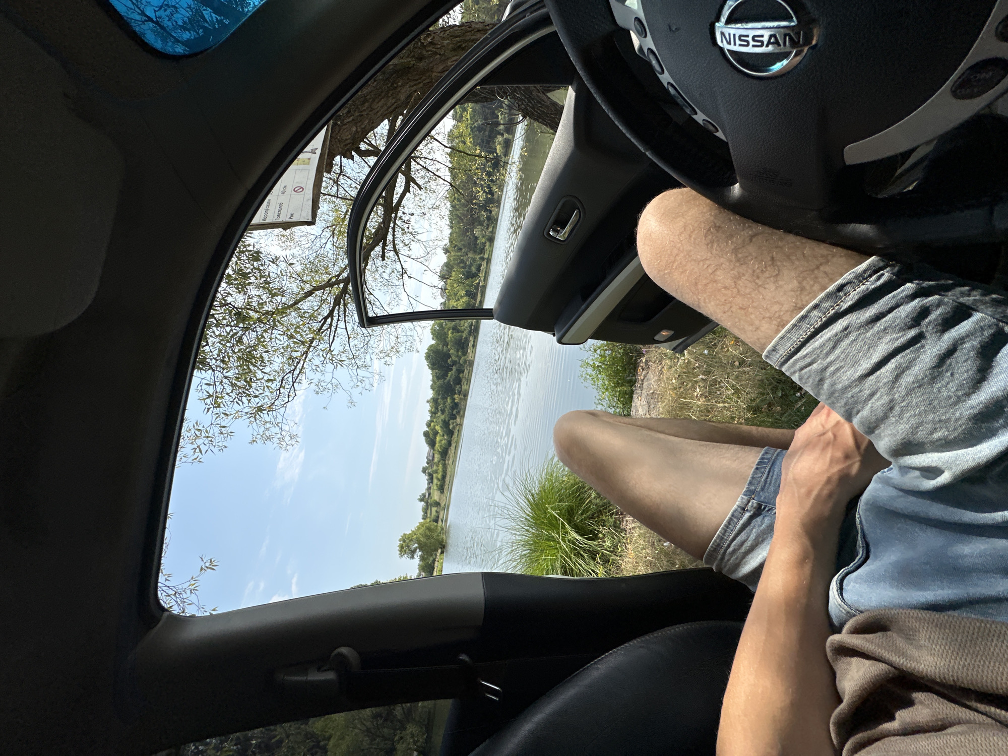 The image shows a first-person perspective from inside a car, parked near a body of water. The car's driver-side door is open, revealing a view of a lake surrounded by greenery. A person's legs, clad in denim shorts, are visible extending out of the car. The steering wheel of the car, with a Nissan logo, is in the foreground on the right. A sign with Cyrillic text is partially visible on a tree to the right of the lake. The sky is blue with scattered clouds. The location is Makiv, Ukraine.