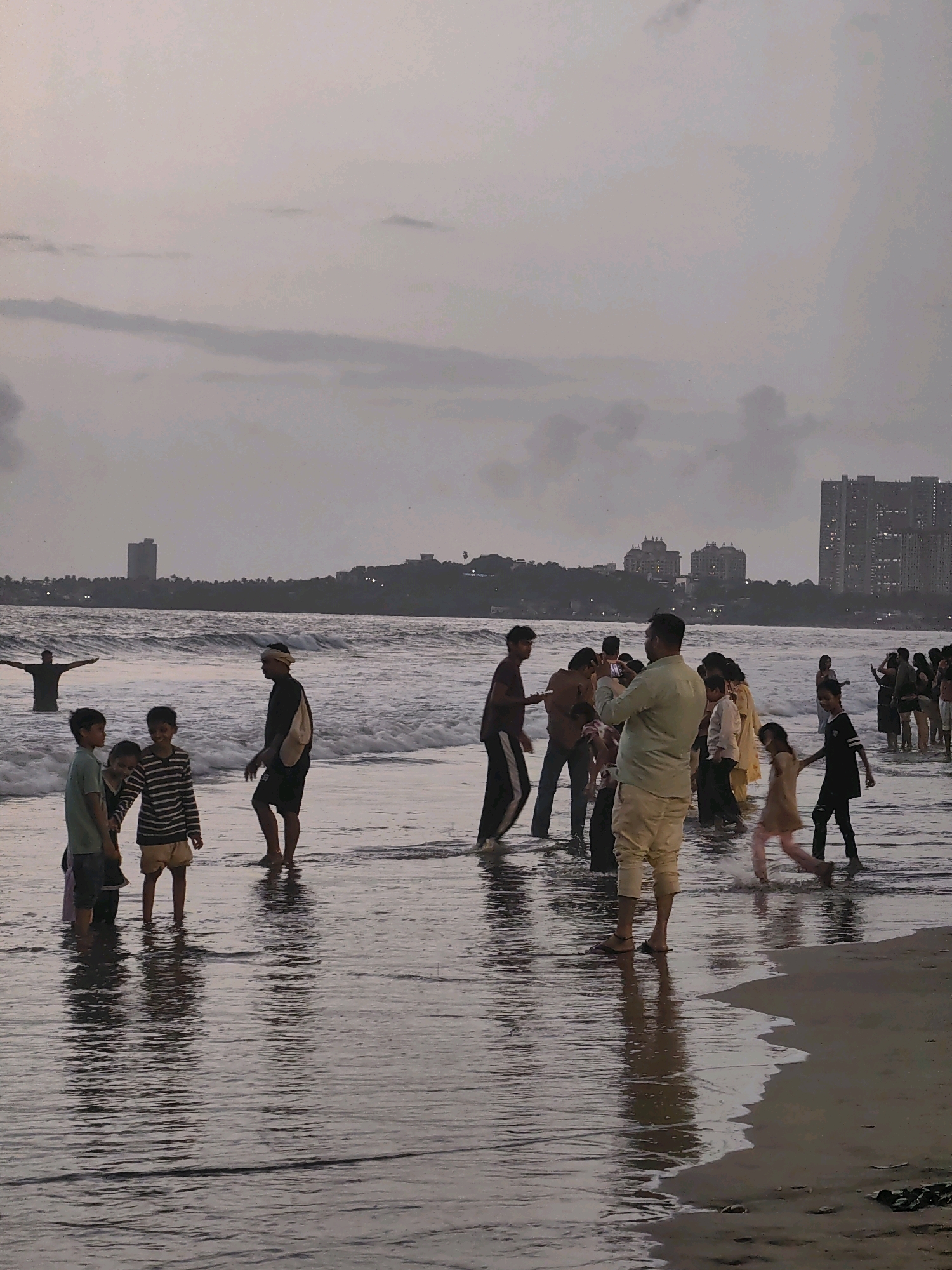 The image depicts a crowd of people, including children and adults, at Juhu Beach in Mumbai, India. The time of day appears to be dusk or early evening, with a cloudy, overcast sky. The sea is moderately wavy, with people wading in the shallow water. In the background, the Mumbai skyline is visible, with several tall buildings and apartment complexes. Some individuals are interacting with each other, with a few taking photographs. The overall scene is one of a typical beach gathering, with a mix of leisure and social activities.
