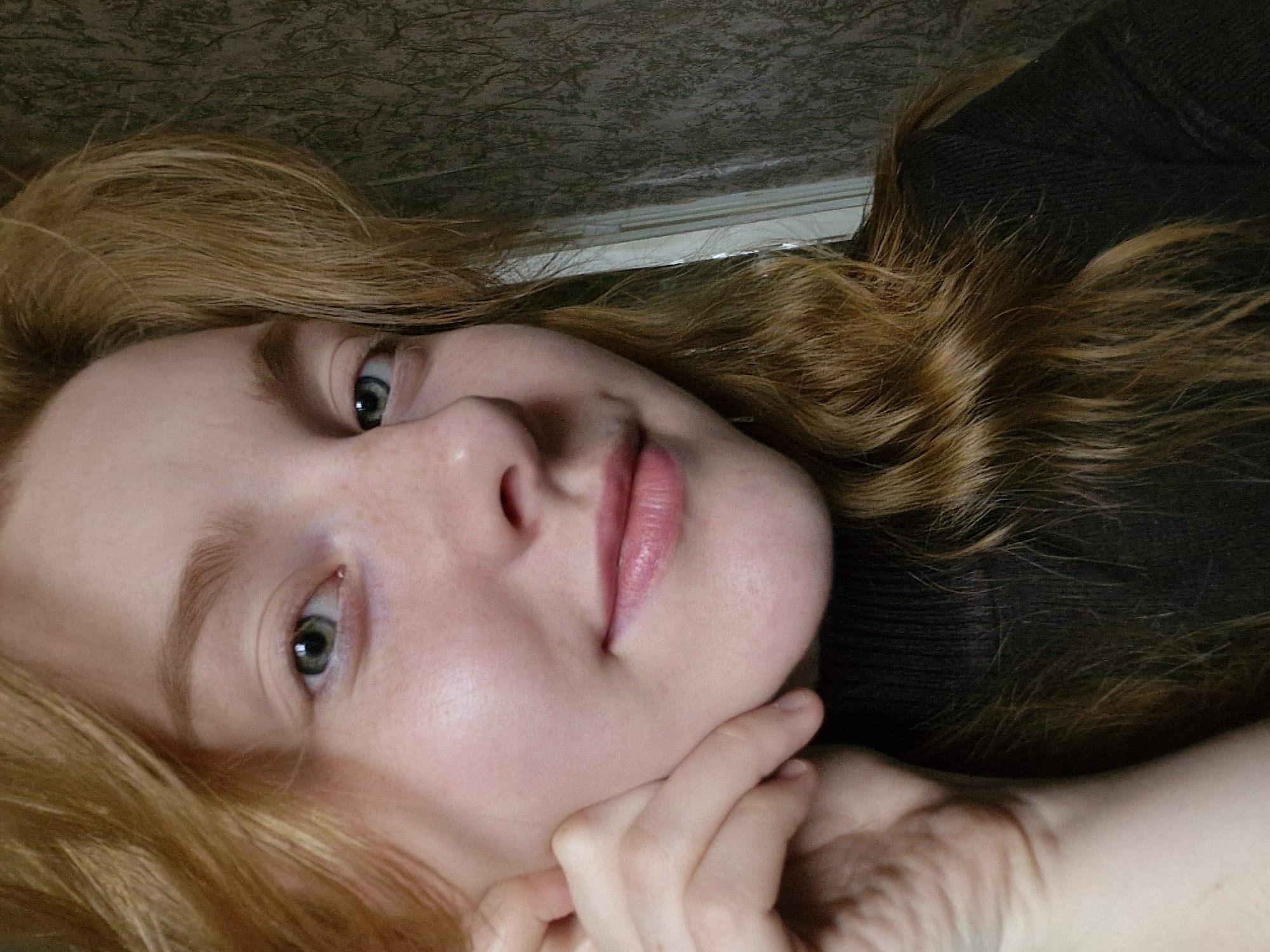 A close-up, sideways portrait captures a young woman with fair skin, light freckles on her nose and cheeks, and long, wavy, reddish-blonde hair. Her eyes, a striking green or hazel, gaze directly at the viewer with a soft, gentle expression, her lips slightly parted. She is wearing a dark, possibly black or dark brown, knitted sweater. Her left hand is subtly placed beneath her chin, drawing attention to her face.
The background suggests an indoor setting, likely a room in Yekaterinburg, Russia, as per the provided context. Above her head is a textured wall or wallpaper, dark green or brownish with a lighter, intricate pattern. Below this, a horizontal band of white, resembling a window frame or baseboard, is visible. The lighting is soft and diffused, creating subtle shadows and highlighting her features without revealing a specific time of day. No text is visible in the image. The overall impression is an intimate and calm portrait.
