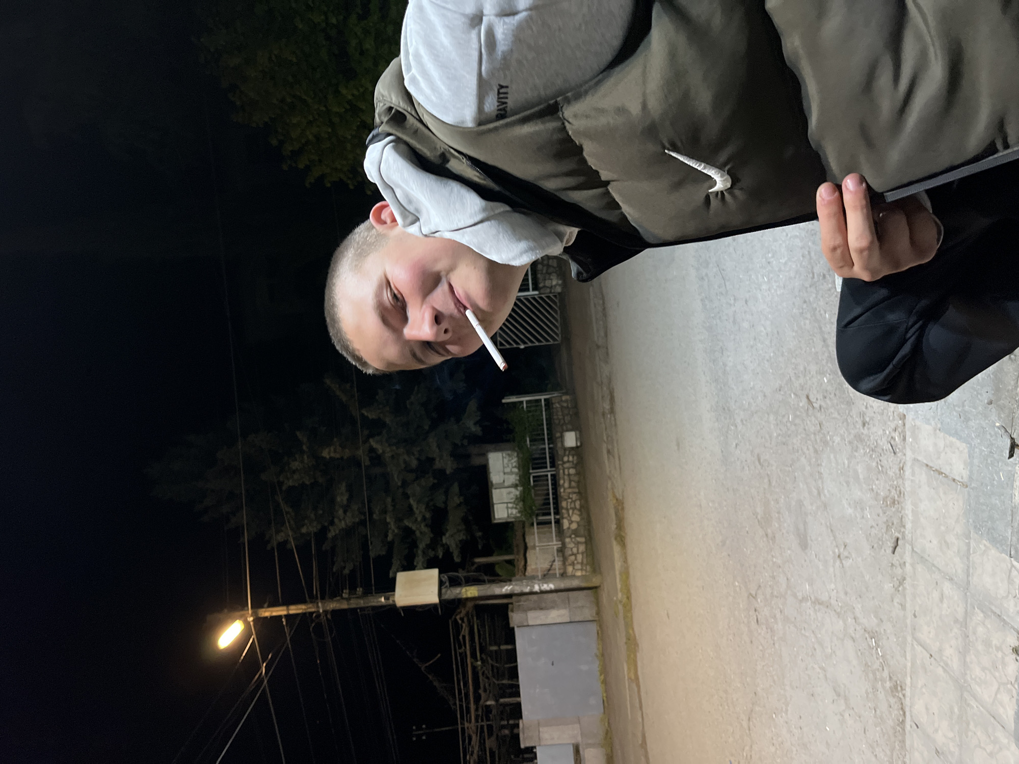 A young man stands on a paved surface in what appears to be a street or sidewalk. He is wearing a grey hoodie under a dark green puffer vest and black pants. He has a very short haircut and is holding a cigarette in his mouth.
The background is dark, with a street light illuminating a tree and some buildings. A utility pole with lines is also visible. Based on the information provided, the location is Karavelovo, Bulgaria.