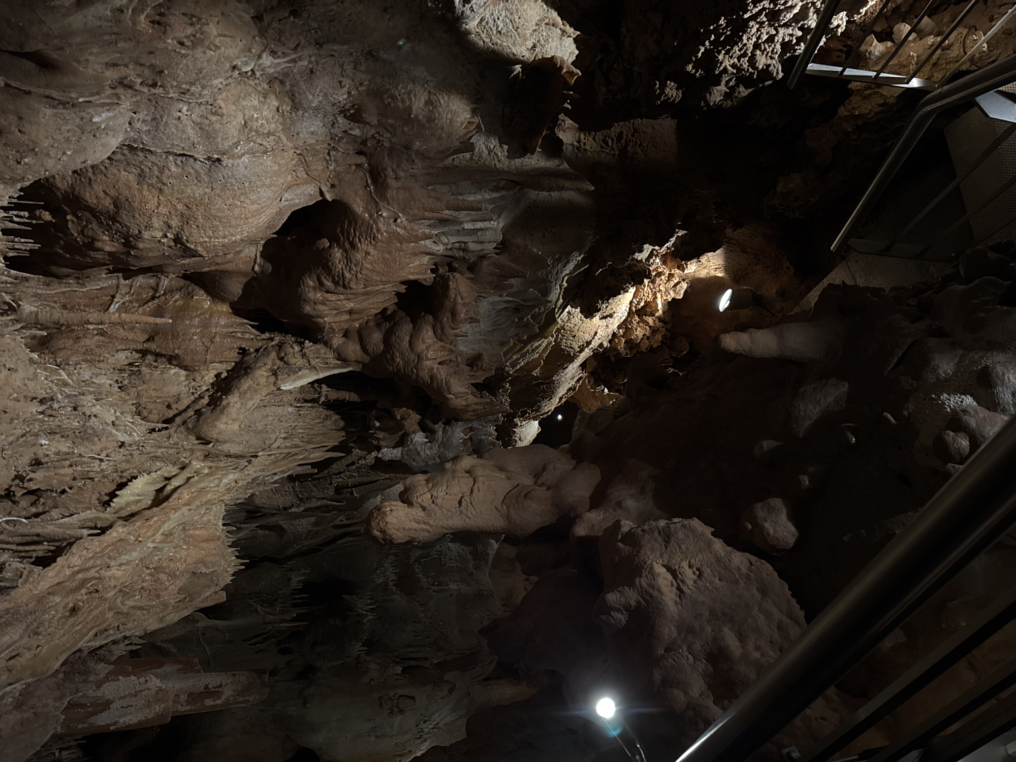 The image shows the interior of the cave of Petralona, located in Πετράλωνα, Greece. The cave features various speleothems, including stalactites and stalagmites, formed from calcium carbonate deposits. Artificial lighting is present, illuminating certain areas of the cave. A metal stairway with a railing is visible on the right side of the image, indicating a pathway for visitors. The lighting and metal features suggest the cave is used for tourism.