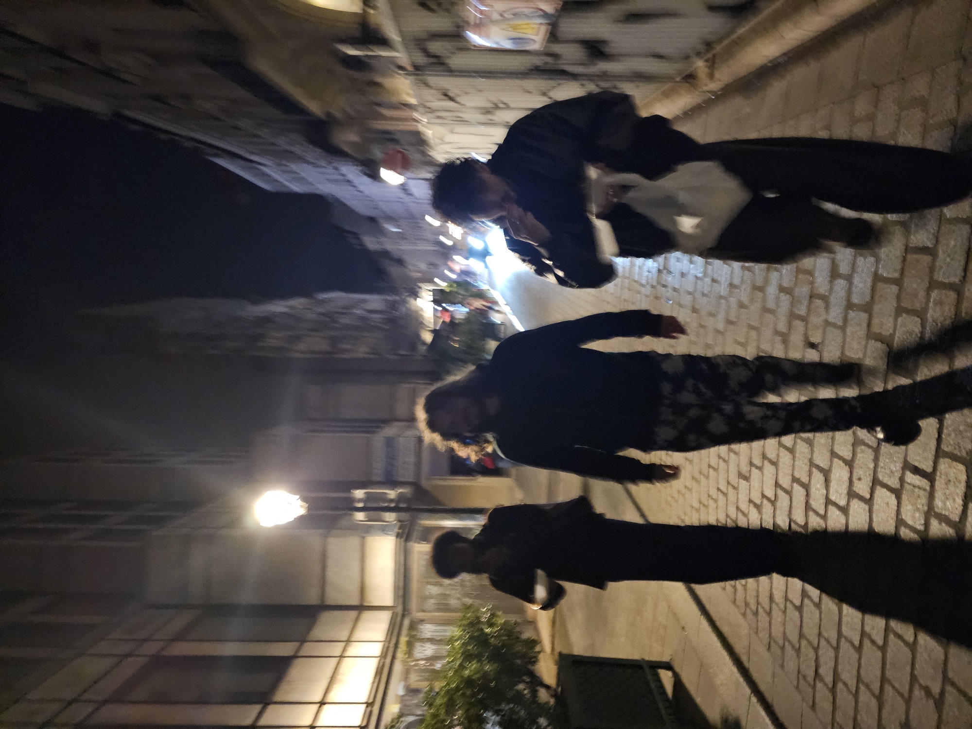 The image depicts an urban scene at night, seemingly in Paris, France. The photo is rotated 90 degrees counter-clockwise, skewing the perspective. Three individuals are present. One individual is at the top of the image, hunched over and holding a white object or bag. A second person is situated in the middle of the frame, their outfit dark. The third individual is at the bottom of the image and appears as a dark silhouette, casting a long shadow onto the cobblestone street. The street is illuminated by a street lamp on the left. Buildings line the left side of the image, while the right side shows the cobblestone pavement. In the distance, there are out-of-focus lights. A small tree or shrub is positioned near the bottom right corner of the photo. The overall environment suggests a narrow Parisian street at night.