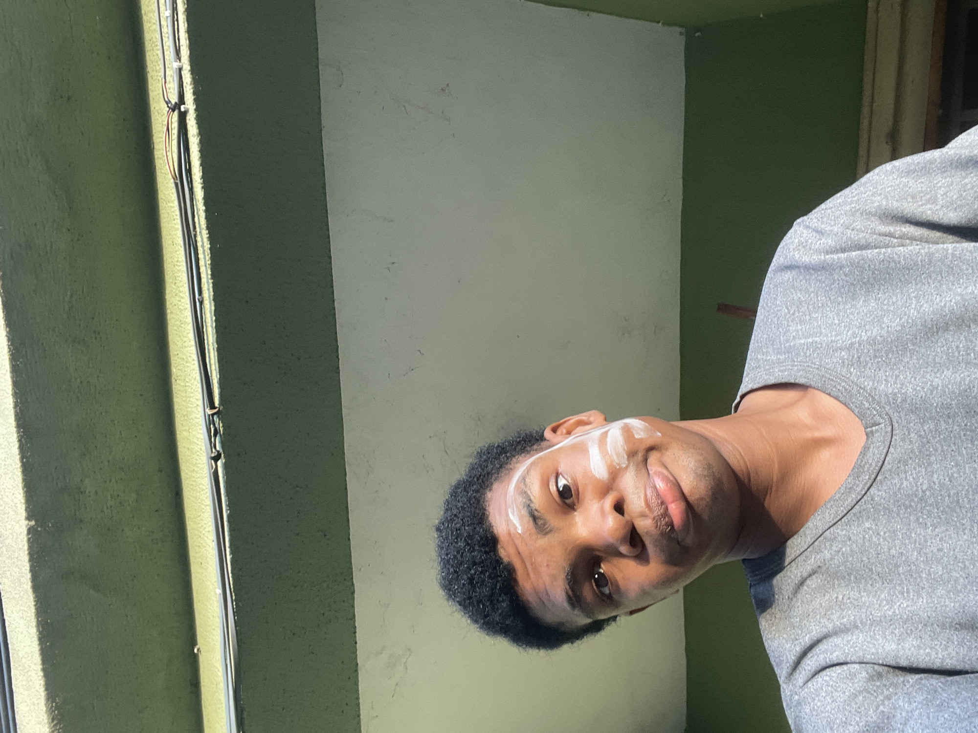 A medium shot of a person with dark skin and short black hair wearing a gray T-shirt. The person is facing the camera and has white markings on their forehead and cheek. The background consists of a vertical section of a green wall on the left, a gray-white wall in the center, and green wall on the right. Electrical wires are affixed to the green wall on the left. The location is most likely in Ado, Nigeria.