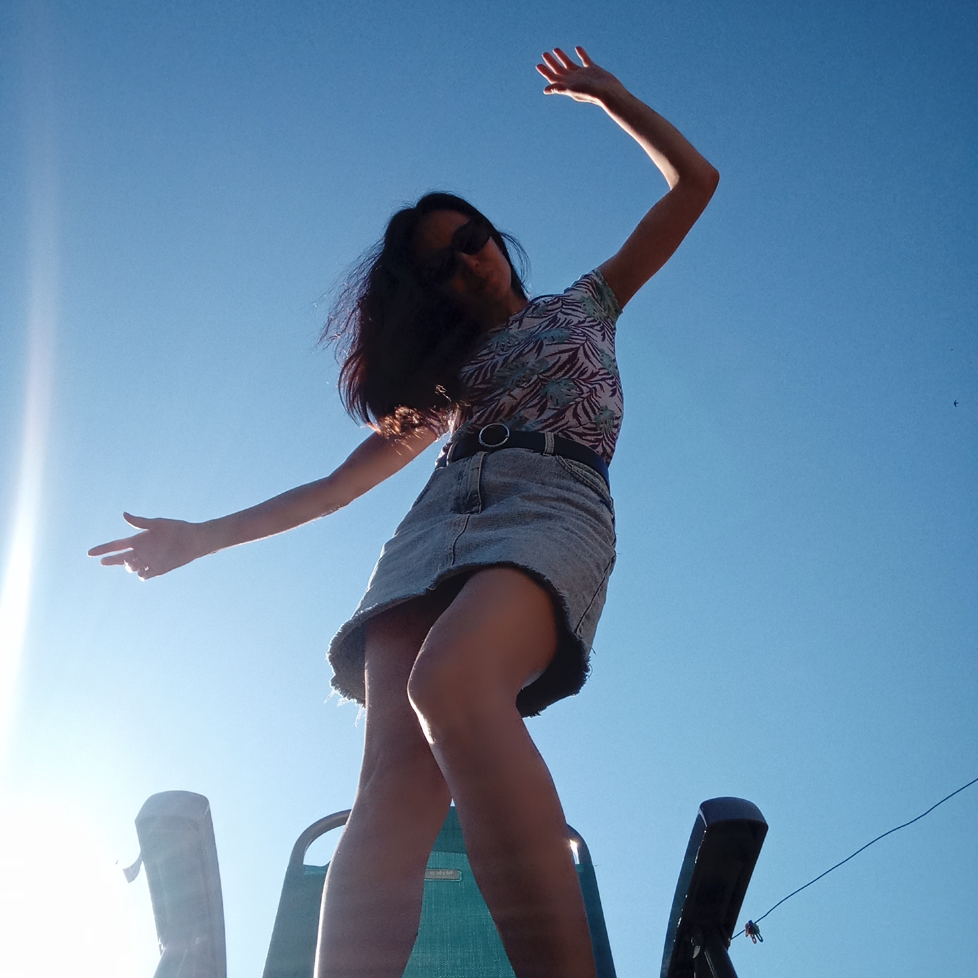 A low-angle shot depicts a woman with long, dark hair wearing sunglasses, a floral patterned shirt, and a denim skirt with a belt. She stands on a green and black lawn chair, arms outstretched, against a bright blue sky with a lens flare on the left. The background features a clear sky with what appears to be a powerline in the upper right corner. The image does not provide enough information to pinpoint a specific location within the city of Буренка, Russia.