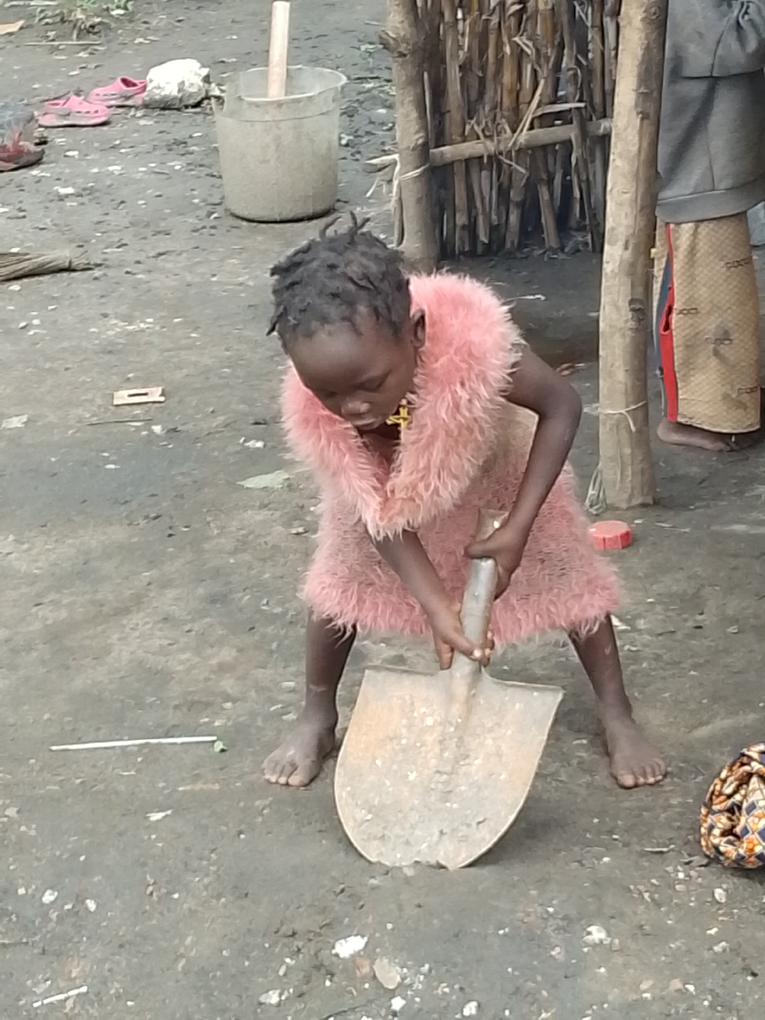 In Otulu, Nigeria, a young Black child is engaged in digging with a shovel. The child is wearing a fluffy pink dress and appears to be focused on the task at hand. The ground is made of dirt and small stones, suggesting an outdoor, possibly rural or semi-urban, setting. In the background, there is a structure made of wood and bamboo, possibly a fence or part of a dwelling. Nearby, a plastic bucket with a wooden stick inside and a pair of pink sandals are visible, hinting at everyday life activities. The lighting suggests it is daytime.